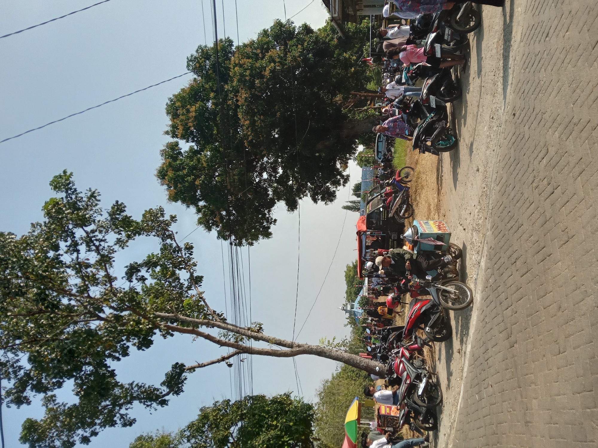 The image depicts an outdoor scene in Ngrambe, Indonesia, on a sunny day. The foreground features a paved area with several parked motorcycles, suggesting a public gathering or market. A red and white stall with a colorful umbrella is visible on the left, with people around it. Further back, more motorcycles are parked, and a crowd of people are milling about. In the background, a large tree with dense foliage dominates the landscape, and another smaller tree is to its left. Power lines are strung across the sky. A blue-domed building can be seen in the distance, possibly a mosque. The overall atmosphere appears to be that of a bustling local event.