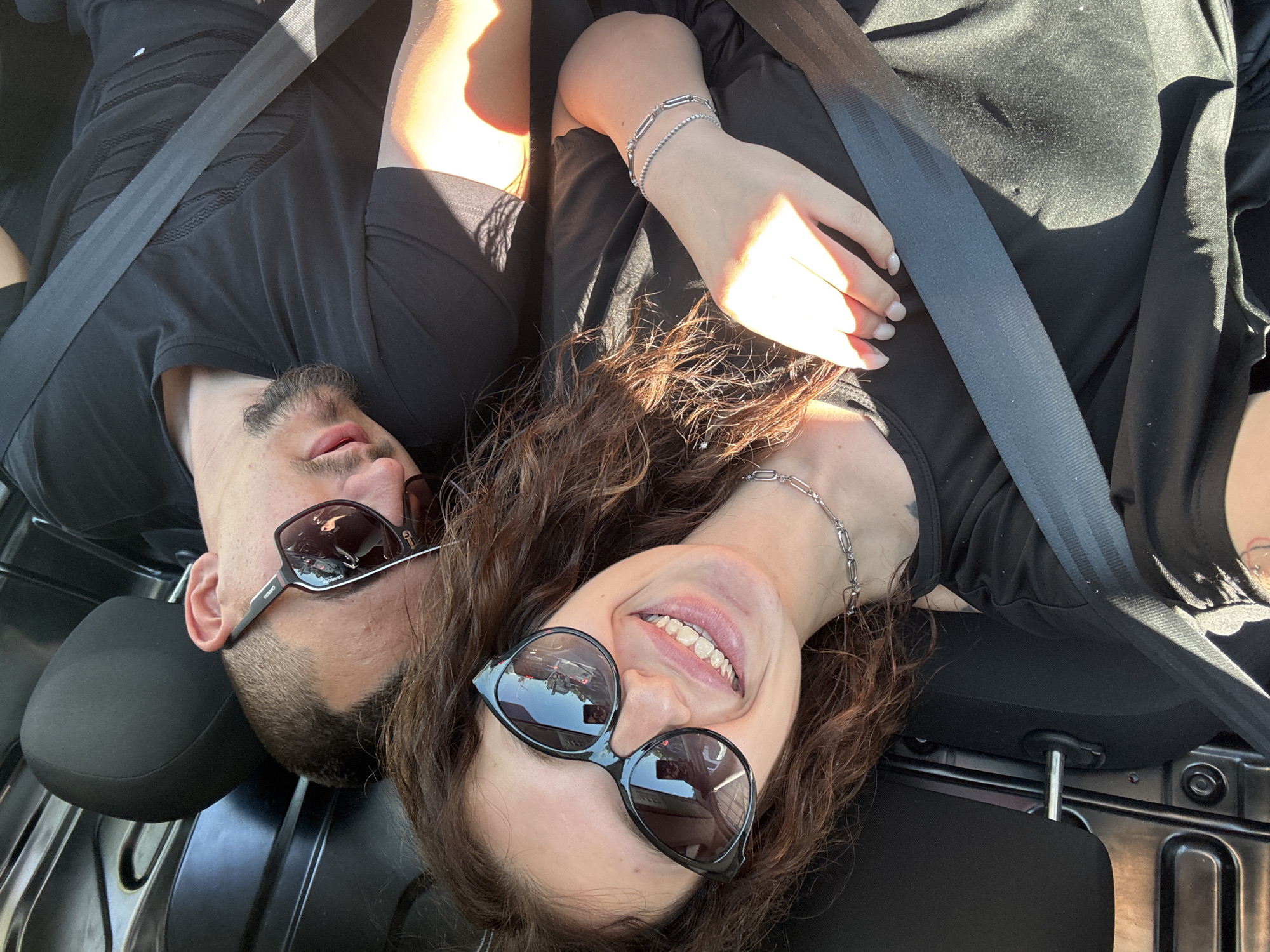 Two individuals are visible in the image, both wearing seatbelts, suggesting they are inside a vehicle. The individual on the left is a male with a beard, wearing sunglasses and a black t-shirt. The individual on the right is a female, also wearing sunglasses, a black t-shirt, and a silver bracelet. She is smiling broadly, revealing her teeth. The reflection in the sunglasses of both individuals hints at a car interior, further supporting the interpretation of the location. The specific location in the city of Unknown, Unknown cannot be determined from the provided image.
