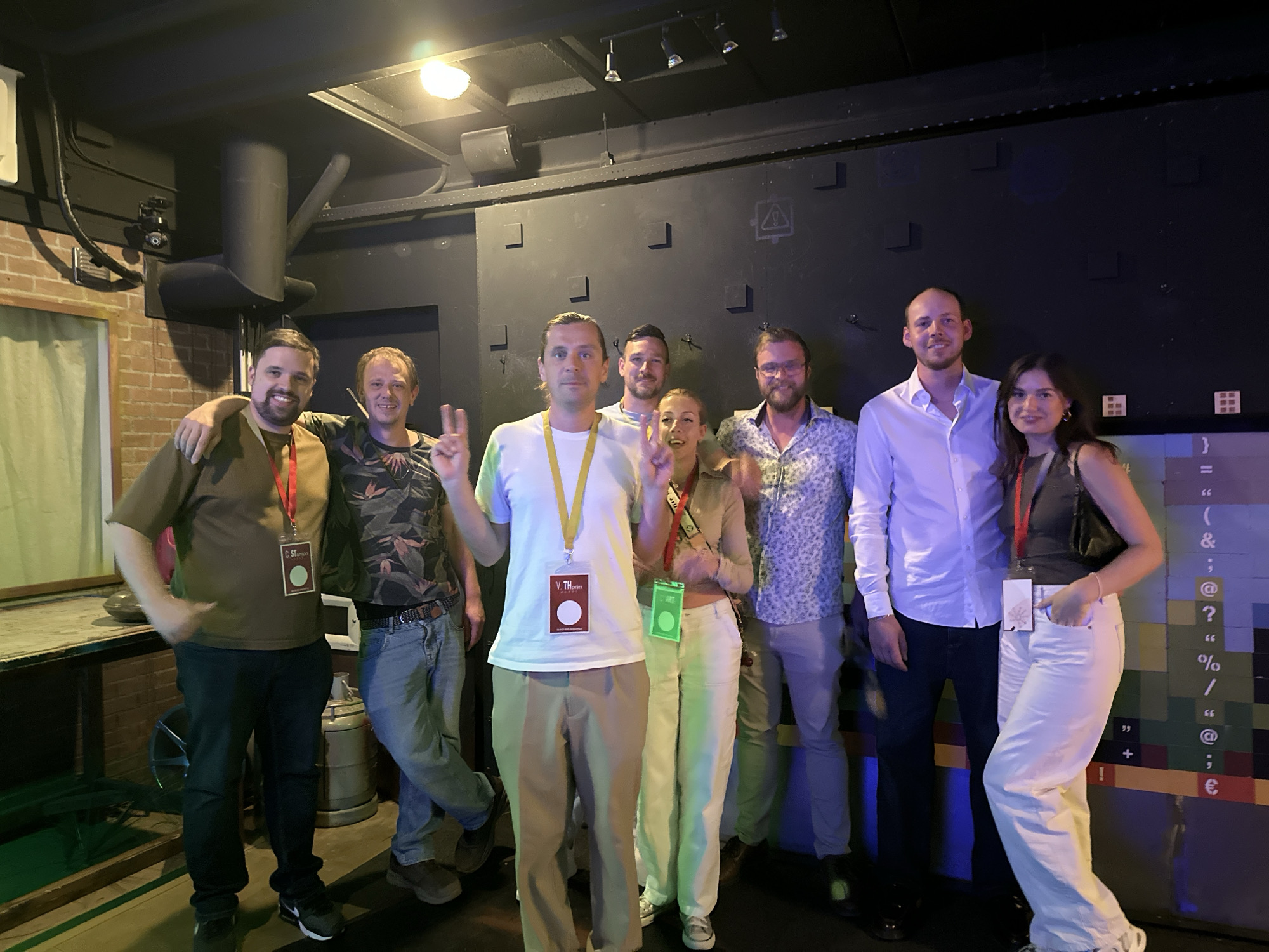 The image captures a group of seven individuals posing for a photograph in what appears to be an indoor venue with dimly lit ambient lighting. The group consists of four men and three women, all of whom are casually dressed. Several individuals are wearing lanyards with identification badges. The background features a dark, textured wall with some abstract graphic elements, and a low-hanging ceiling with exposed industrial-style lighting fixtures. To the left, a brick wall and a wooden surface are visible. The overall atmosphere suggests a social gathering or event. The image is devoid of any specific landmarks or indicators that would pinpoint its location within Amsterdam, Netherlands, or any other city.