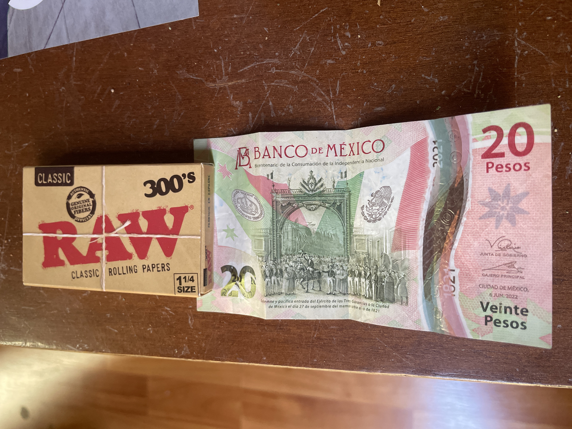 The image shows a box of RAW brand rolling papers, a 20 Mexican Peso bill, and a wooden surface. The rolling papers are a light brown color with red lettering. The 20 Peso bill is green, pink, and white, and shows a historical scene on one side. The wooden surface is a medium brown color.
