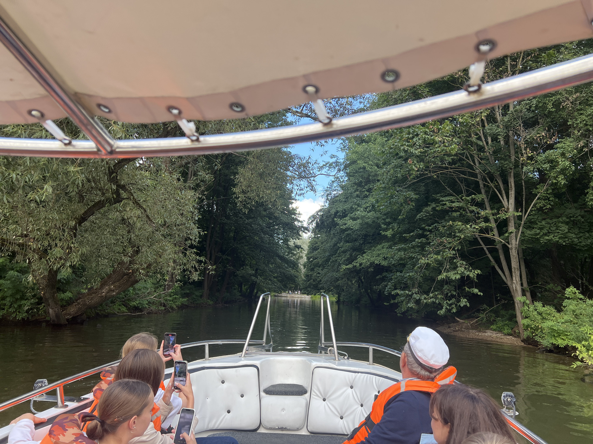 The image shows a boat ride on a calm river, likely in or near Minsk, Belarus. The boat, which is white with silver railings, is moving forward, with a canopy overhead. Several people are visible on the boat, some of whom are holding up phones to take pictures or videos. The riverbanks are lined with lush green trees, creating a tunnel-like effect with the sky visible through the branches. The overall scene suggests a leisurely outing in a natural setting.