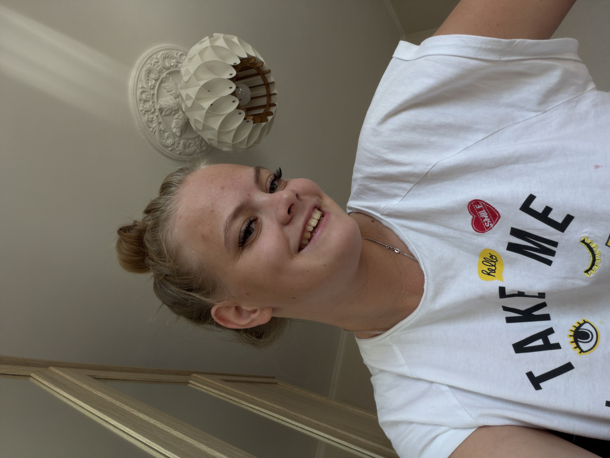 A young woman with blonde hair tied in a bun smiles at the camera, her head tilted to her right. She is wearing a white t-shirt with the words "TAKE ME" printed in large black letters across the front, accompanied by graphics of an eye and a mouth. A small yellow speech bubble with the word "hello" is on the left side of her chest, and a red heart with the word "SMILE" is on the right. She is also wearing a silver necklace. The background shows a wooden structure on the left and a white ceiling fixture with a light bulb on the right. The overall scene is indoors. The location is not discernible from the image.