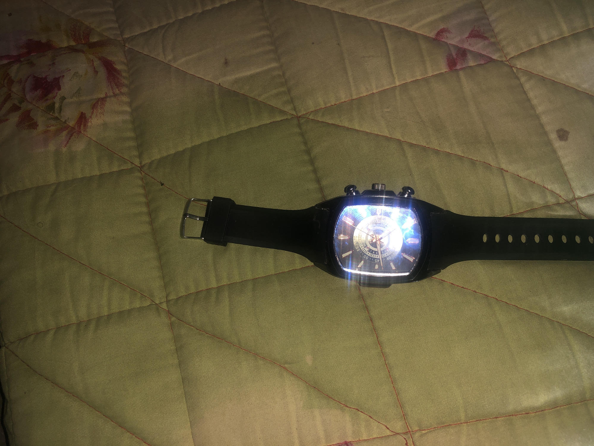 An overhead shot captures a black wristwatch with a luminous dial placed on a quilted surface. The surface is a light green color with a floral pattern, and it appears to be a bedspread or blanket. The watch is oriented vertically, with the strap extending downwards. The watch face is brightly lit, obscuring some of the details, but Roman numerals and markers are visible on the bezel. The background is out of focus, highlighting the watch as the primary subject. No specific location in Jalingo, Nigeria, can be identified from the image.