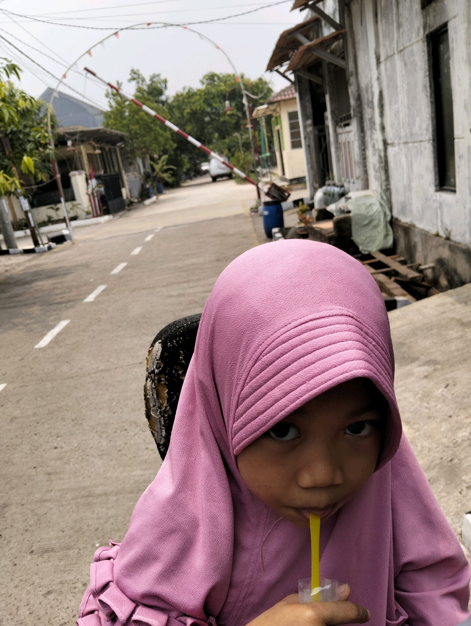 A young person wearing a pink hijab drinks from a yellow straw attached to a plastic cup. The person is positioned in the foreground, with their face partially visible. Behind them, a damaged tire is present. The background depicts a street scene in Cileungsi, Indonesia, with a road marked with white dashed lines, buildings on either side, and a red and white striped barrier in the distance. Power lines crisscross overhead. The sky is overcast.