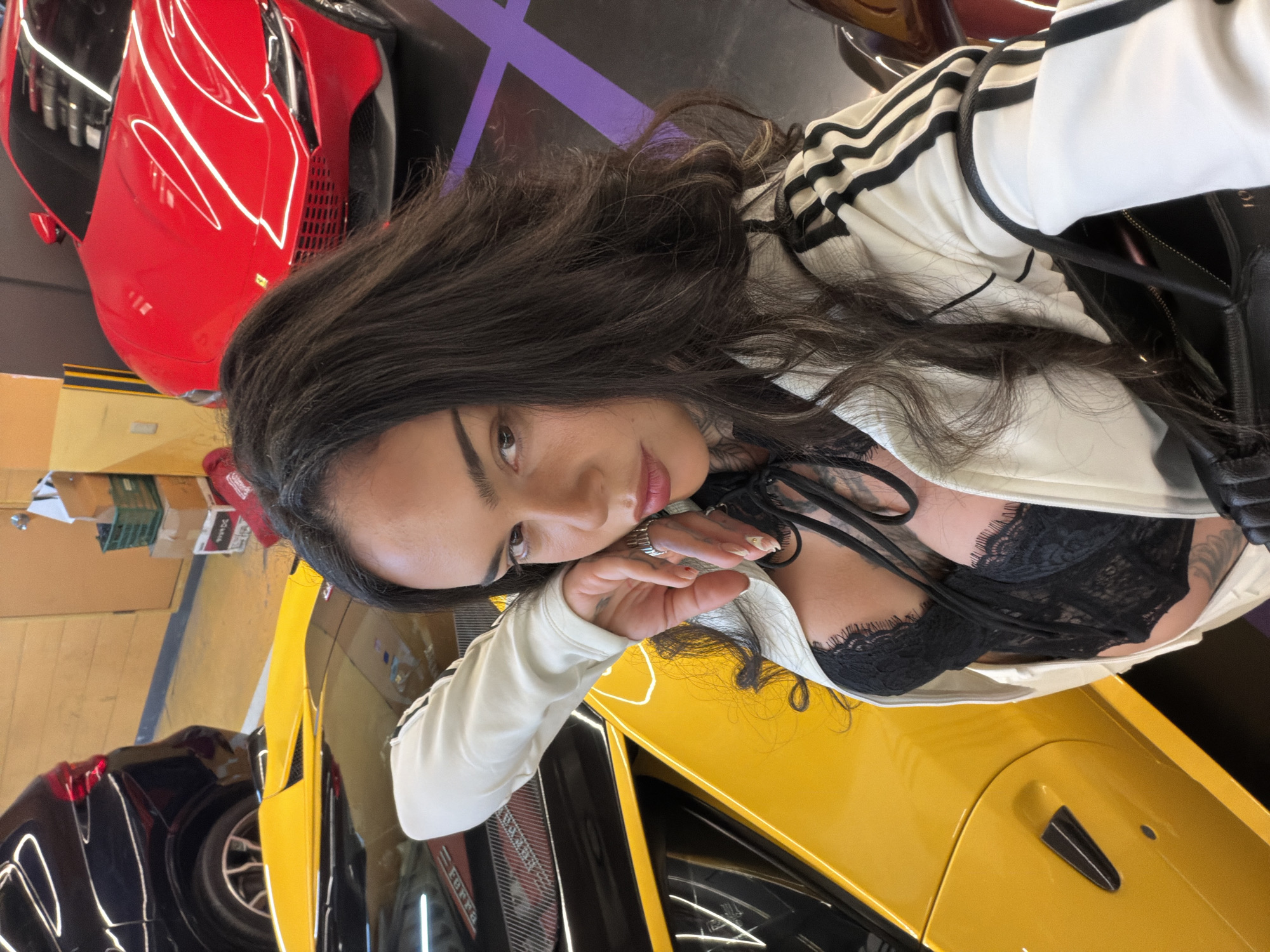 A woman with dark hair, wearing a white Adidas jacket over a black lace top, is posing next to a yellow Ferrari. In the background, there are additional cars, including a red Ferrari and a black car. The setting appears to be a car dealership or showroom. The location is likely Seoul, South Korea.