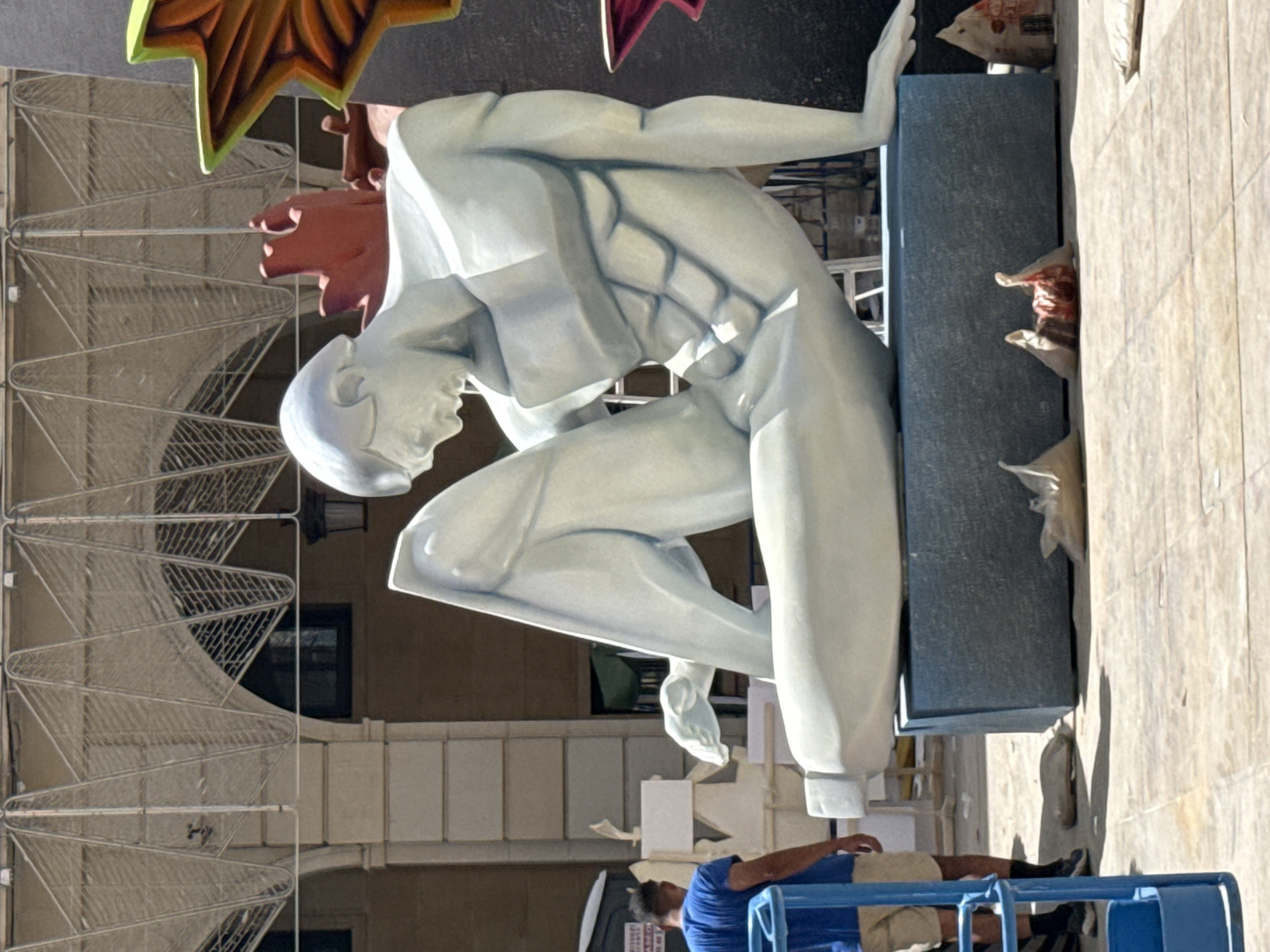 An outdoor, sunlit scene depicts a large, pale blue, abstract sculpture of a reclining male figure. The sculpture is positioned on a dark blue rectangular base. To the left of the sculpture, a man in a blue t-shirt and tan shorts stands facing away from the camera, looking towards the sculpture. He is near a blue metal lift. The background features architectural elements including columns, arches, and a decorative metal structure overhead. Various construction materials and objects, including scaffolding, bags, and small structures, are scattered around the scene.