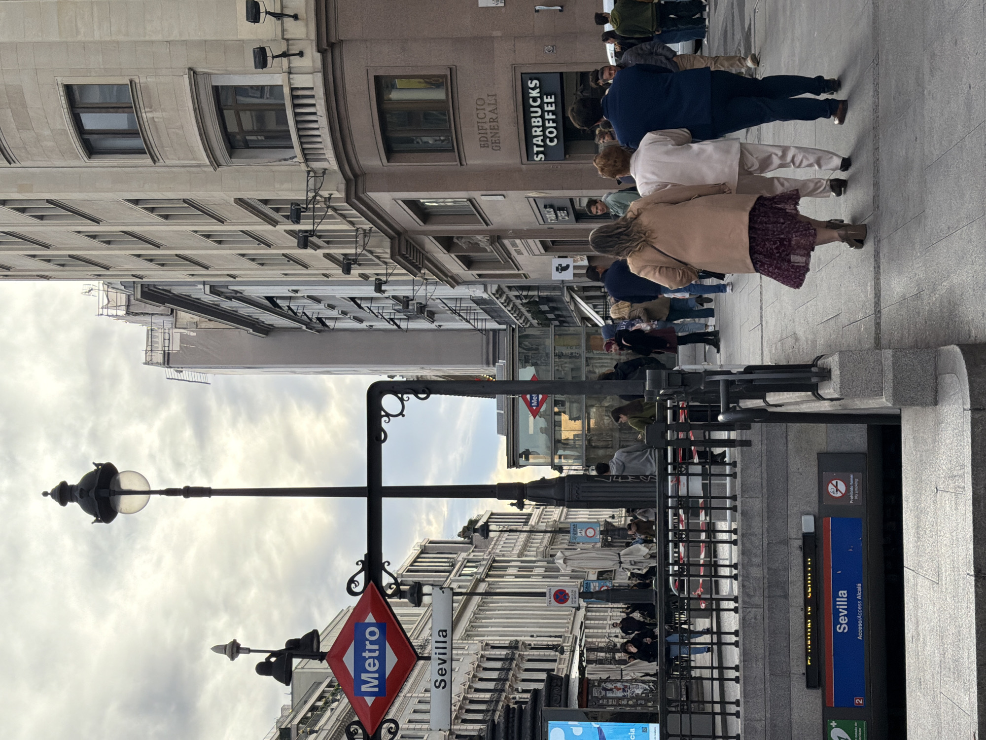 This image depicts the entrance to the Sevilla Metro station in Madrid, Spain. The station entrance is marked by a red diamond logo with "Metro" written in white and a sign reading "Sevilla." People are walking on the sidewalk near the entrance. A Starbucks Coffee is visible in the background with "Edificio Generali" above it. The architecture of the surrounding buildings is typical of Madrid. There are streetlights and various signs present.