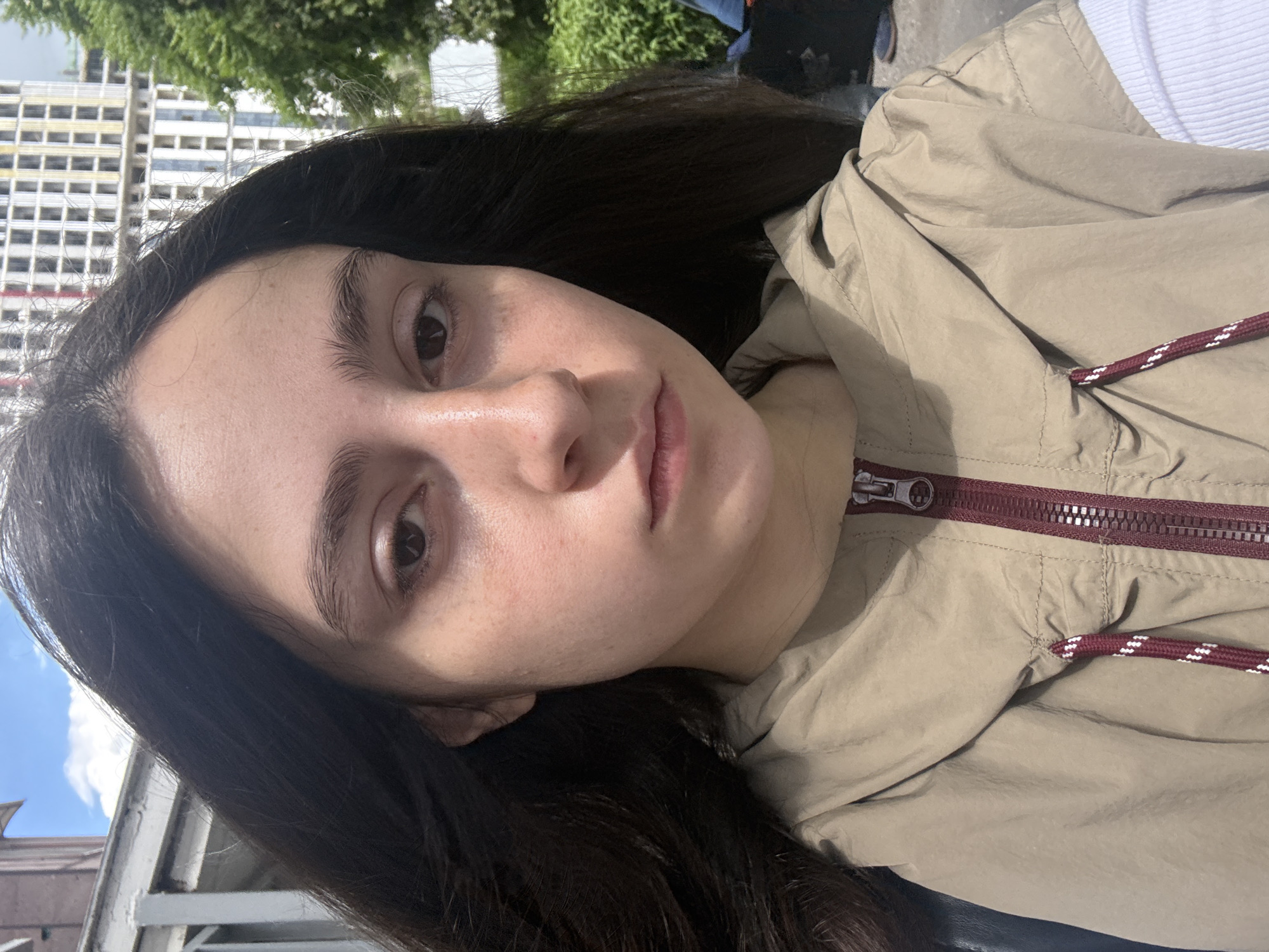 A person with dark hair is facing the camera, wearing a tan jacket with a maroon zipper and drawstring. The background includes a building with repetitive windows and greenery, suggesting an urban environment. The presence of the sky indicates it's daytime. The location is Yerevan, Armenia.