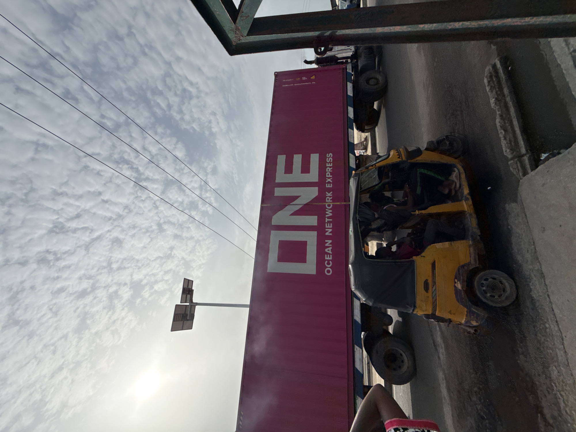 The image shows an urban street scene in Ado, Nigeria. A red shipping container with "ONE OCEAN NETWORK EXPRESS" in white lettering is being transported by a truck. A yellow and black auto-rickshaw (keke) is stopped adjacent to the truck. A few people are visible within the auto-rickshaw. Overhead power lines are present, along with a utility pole. The sky is overcast. A section of a building is on the right.