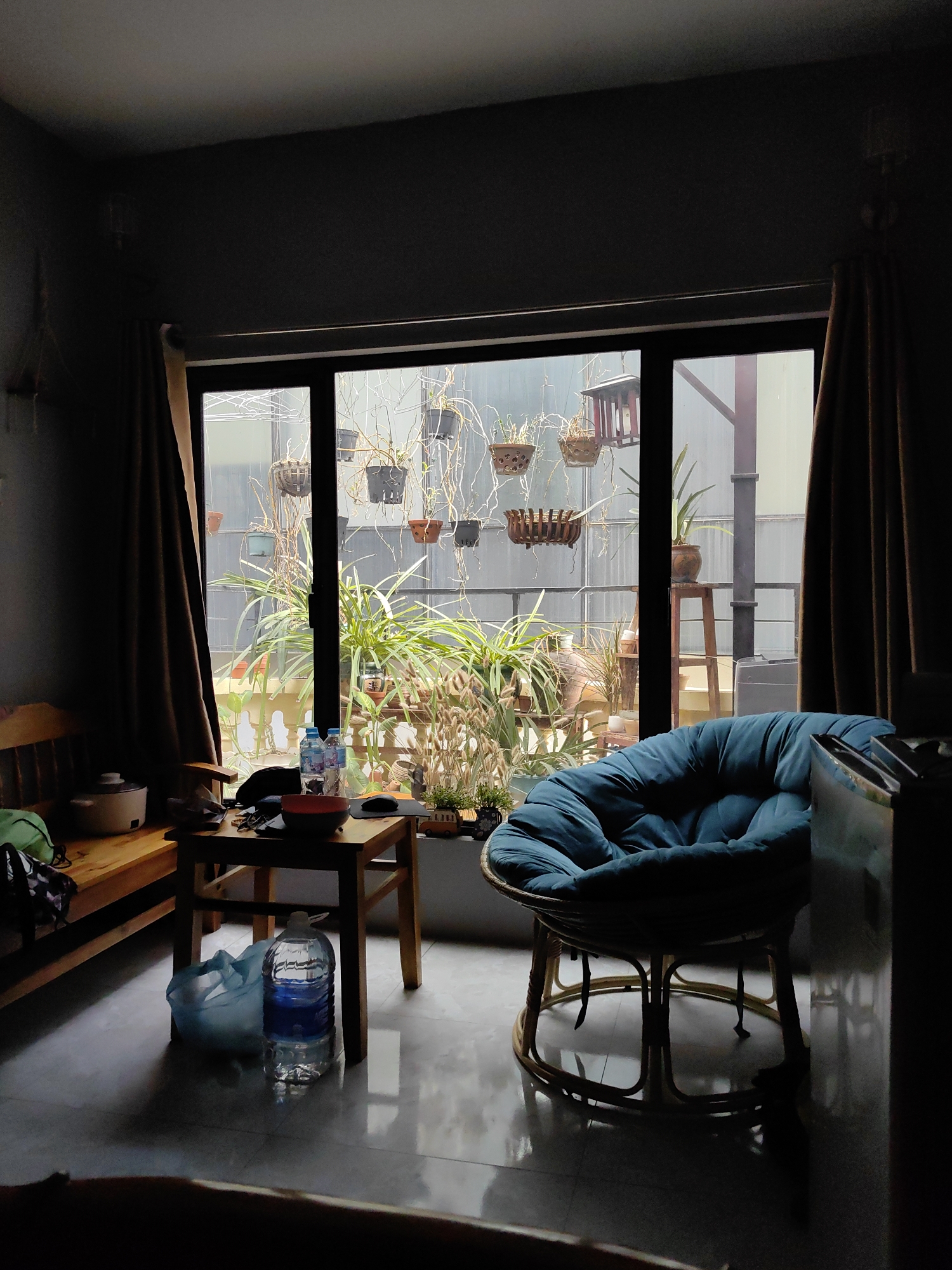 The image depicts an interior scene in Hà Nội, Vietnam, characterized by a large window overlooking an exterior area with plants. The room is dimly lit, with the primary light source being the daylight filtering through the window. The window is flanked by heavy brown curtains. Outside, various potted plants are suspended from above, creating a cluttered yet naturalistic view. Below, more plants are arranged on a surface near the window. Inside the room, a wooden bench sits to the left, accompanied by a small wooden table cluttered with objects, including water bottles, a bowl, and other miscellaneous items. On the right, there is a blue papasan chair. A large bottle of water and a plastic bag sit on the floor. The overall scene is one of domesticity, albeit somewhat disorganized, and showcases the integration of indoor and outdoor spaces common in urban environments.
