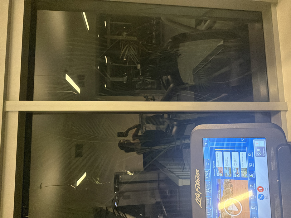 A window displays reflections of gym equipment, lighting, and a person holding a phone. A Life Fitness treadmill screen is prominently visible in the lower right corner. The screen displays a workout program, distance, time, calories, and pace information. Several workout options are shown on the screen with imagery of landscapes such as Auckland and the South Island. The treadmill speed is set to 3.5.