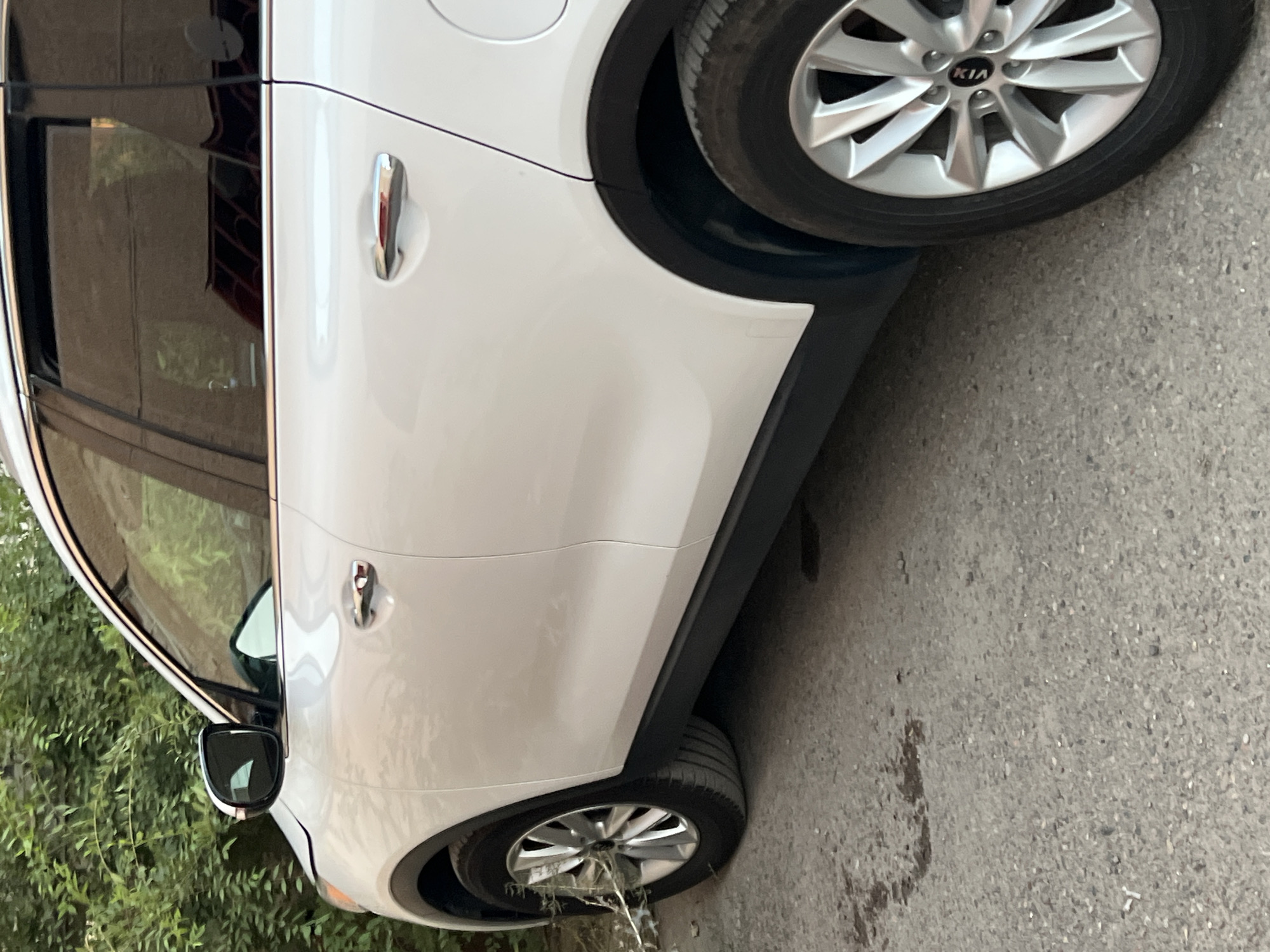 A white Kia Sorento SUV is parked on a textured asphalt surface. The vehicle is seen from its right side, with the front passenger door and rear passenger door visible. The car has chrome door handles and a black trim along the lower body and wheel arches. The front wheel is a silver alloy with the Kia logo in the center. Behind the car, dense green foliage is visible, suggesting it is parked near trees or bushes. A brick wall is reflected in the car's windows. The image appears to be taken in an outdoor setting, likely a residential area or a parking lot in Tokmok City, Kyrgyzstan, given the context. There are no people visible in the image.