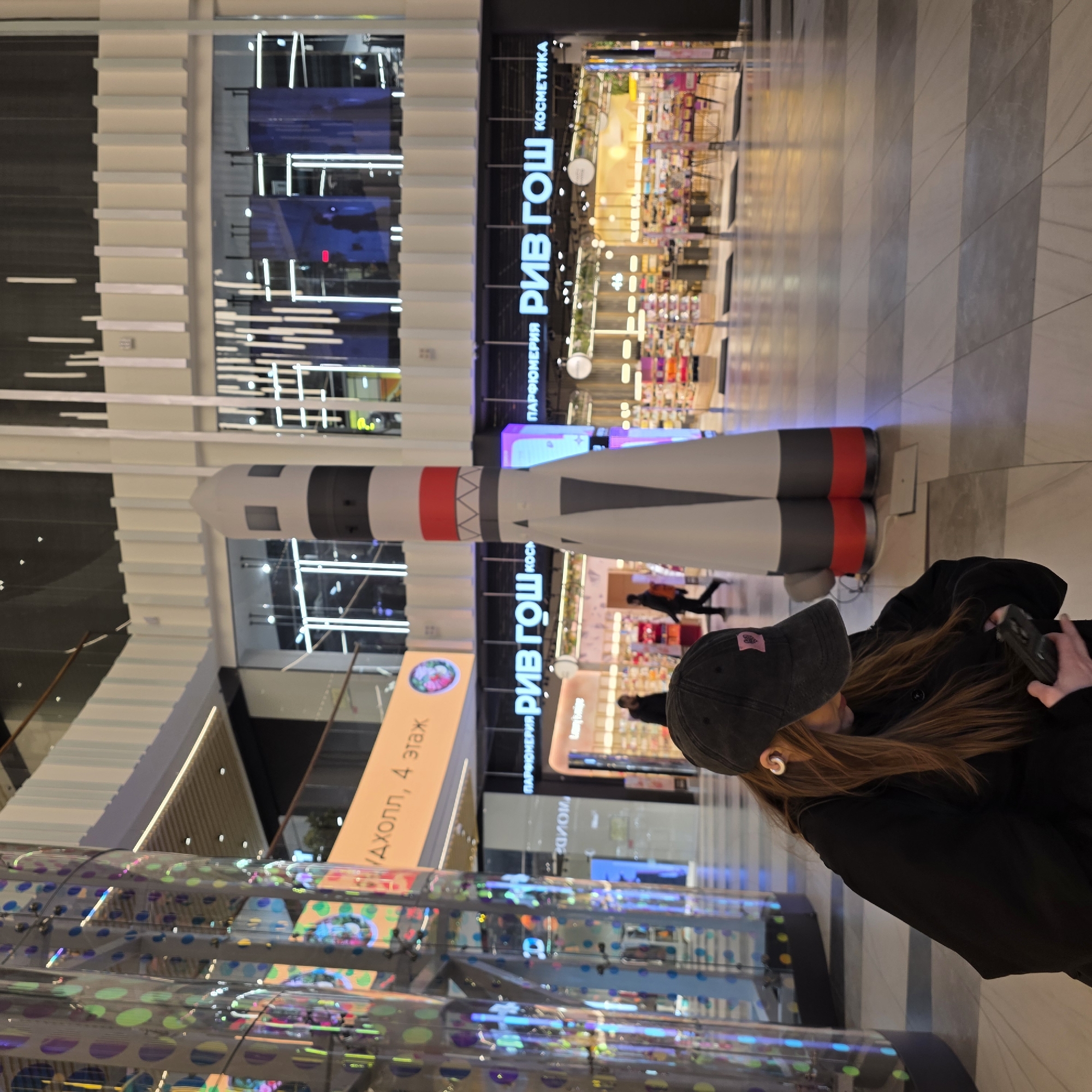 A vertical indoor shot presents a scene at the Galeria shopping center in Saint Petersburg, Russia. A person with long brown hair, wearing a black baseball cap and jacket, appears to be taking a photo with a phone. To the left of the person is a transparent structure decorated with colored dots. In the background stands a model of a rocket with red and gray segments, positioned in front of several storefronts, including "РИВ ГОШ" (RIV GOSH), a Russian chain of perfumery and cosmetics stores. Other signs and displays are visible in the distance, along with the typical flooring and architectural details of a large shopping mall.