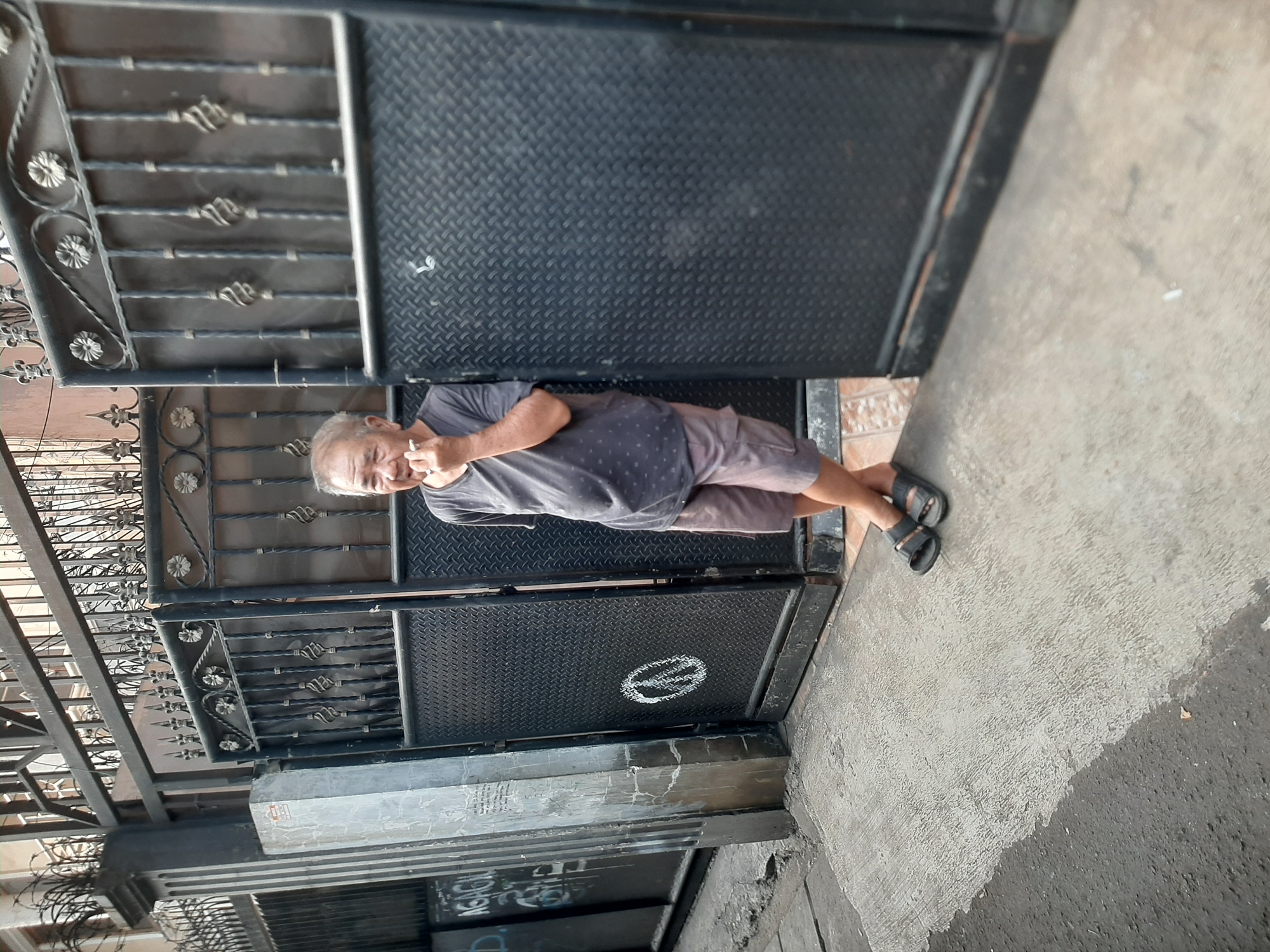 An elderly Asian man, with graying hair and wearing a dark patterned t-shirt and light brown shorts, stands with his legs crossed and leans against a metal gate in an urban setting. He holds a cigarette to his mouth with his right hand. The gate is black, ornate at the top, and has diamond-patterned metal panels. To the left of the gate, a concrete pillar with some graffiti is visible. The ground is paved with concrete and asphalt. A peace sign is visible on one of the gate panels. The scene appears to be in a residential area of Central Jakarta, Indonesia.