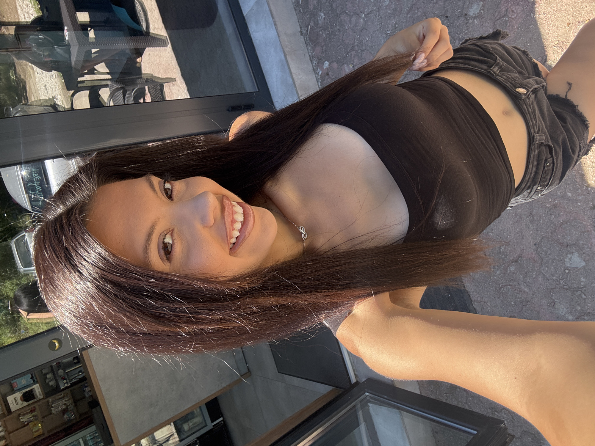 A woman with long, dark hair is captured in a close-up, vertically oriented selfie. She is wearing a black, strapless top and distressed black denim shorts. Her left arm is extended towards the camera, and her right hand is in her hair, near her face. She has a bright smile, showing her teeth, and appears to be looking at the camera. A delicate silver necklace with an infinity symbol pendant is visible around her neck.
The scene suggests an outdoor setting, with a concrete-like ground and dappled sunlight illuminating the woman's skin. In the background, partially visible through reflections in what appears to be a glass door or window, are outdoor furniture, possibly tables and chairs, and some greenery, suggesting a patio or dining area. Further in the distance, a building interior with shelves stocked with items is discernible through the glass. The lighting indicates it is daytime, likely sunny. The overall mood is casual and happy. The location context provided is Sofia, Bulgaria. There is no visible text in the image.