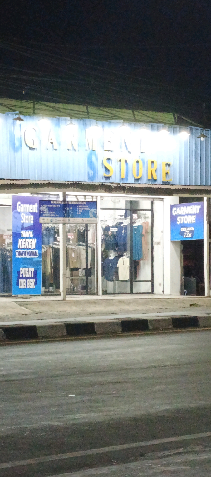 This media file shows a night-time exterior view of a "Garment Store" located in Banyuraden, Indonesia. The store's facade features prominent signage with "GARMENT STORE" written in large, yellow, three-dimensional letters against a light blue background. The store appears to be a retail establishment selling clothing.
Inside the store, which is visible through large glass windows, racks of clothing can be seen. Some of the garments are displayed on mannequins or hangers, including what appear to be shirts and dresses in various colors like blue, teal, and cream.
In front of the store, to the left, there is a large blue banner with white text that reads "Garment Store" at the top. Below this, it says "TAMPIL KEREN TANPA MAHAL" which translates to "Look Cool Without Being Expensive." Further down the banner, it states "PUSAT IDR 85K," indicating it's a center for items priced at 85,000 Indonesian Rupiah. On the right side of the entrance, another blue sign with white text also reads "GARMENT STORE" and displays "DISKON 12K," suggesting a discount of 12,000 Rupiah.
The scene is illuminated by artificial lighting, typical for a commercial area at night. The street in front of the store is paved and has a sidewalk with a concrete curb. The overall impression is of a busy commercial street at night, with a focus on a clothing store advertising affordable fashion. There are no people visible in the image, and the weather appears clear.