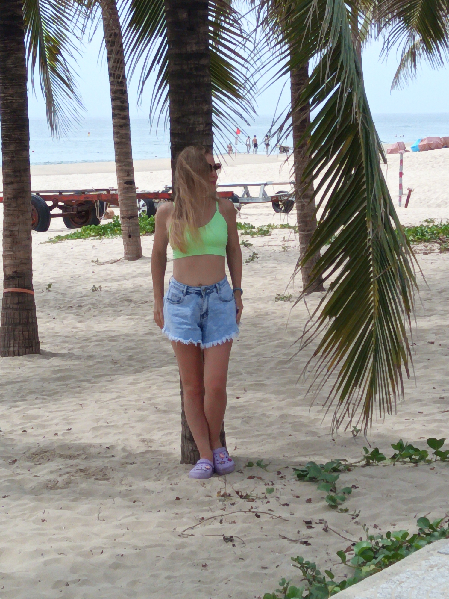 The image depicts a woman standing on a sandy beach in Da Nang, Vietnam, with palm trees in the foreground. She is wearing a bright green bikini top and denim shorts. Her legs are crossed, and she is leaning against a palm tree. In the background, the ocean is visible, with a few people walking along the shore and some structures that appear to be related to beach activities. The sand is light in color and appears to be a typical beach environment. There is a trailer with wheels visible to the left of the woman, likely used for transporting boats or equipment.