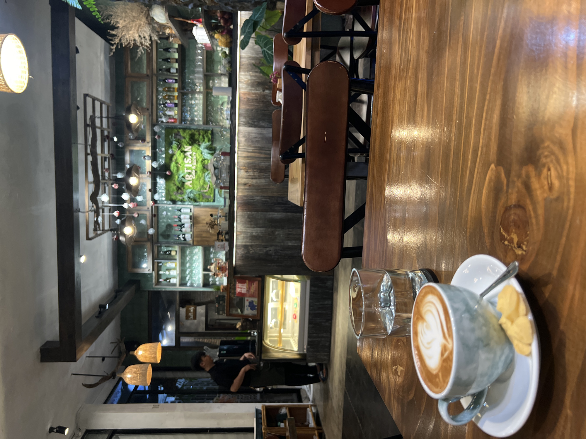 The image captures the interior of a cozy cafe or eatery, likely in the evening given the reliance on artificial lighting. The perspective is from a wooden table, looking towards the back of the establishment.
In the foreground, a light-colored cup of coffee with latte art is placed on a white saucer, accompanied by a small spoon and a few pieces of what appear to be shortbread cookies. A clear glass is also on the dark wooden table, reflecting light. The table itself has a prominent wood grain, suggesting a rustic aesthetic.
The midground features a row of empty wooden stools with brown seats aligned along a bar or counter. Further back, a display case with illuminated shelves showcases desserts or other items. To the left of the display case, a person in a dark uniform, possibly an employee, is standing and appears to be attending to something behind the counter.
The background reveals a well-decorated space with various shelves stocked with bottles, possibly wine or spirits, implying a bar component to the establishment. A striking green wall art piece with moss and foliage stands out, and partially visible text on it reads "ARTISAN Cafe & Eatery." The lighting consists of various fixtures, including woven pendant lamps and decorative lights hanging from the ceiling. One sign partially visible near the counter reads "Self only." The overall ambiance is warm and inviting, characterized by wood textures and soft lighting.