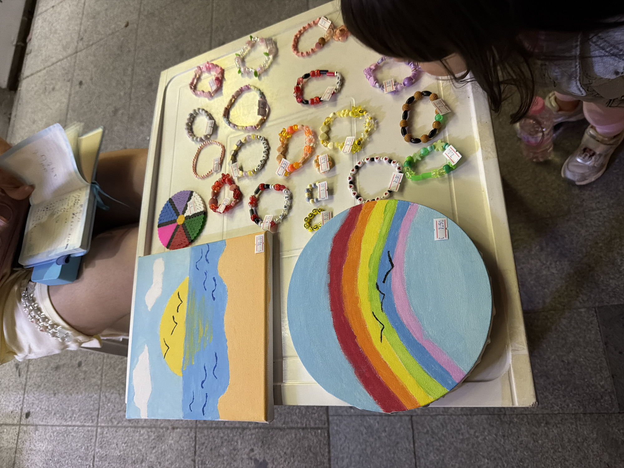 A young girl with long dark hair is sitting on the ground, facing away from the camera, looking at a table laden with handmade bracelets and two small paintings. The table is white and appears to be a portable display.
On the table, to the left, are two paintings. The top one is a small canvas depicting a sunrise over the ocean with three birds flying in the sky. The bottom painting is larger and circular, featuring a vibrant rainbow with three black birds flying across it. Both paintings have price tags.
Arranged on the table are numerous colorful bracelets, mostly made of beads. Some bracelets have letters that spell out words, while others are adorned with charms like shells and flowers. Each bracelet also has a price tag.
To the right of the paintings and bracelets, a person's legs and torso are visible, wearing a white dress with pearl embellishments around the waist. This person is holding an open book with delicate, lace-like pages.
The table is positioned outdoors on a cobblestone surface, suggesting it might be a street market or sidewalk display. The lighting indicates it is daytime. The scene is likely in Burgas, Bulgaria, given the context of the request.
