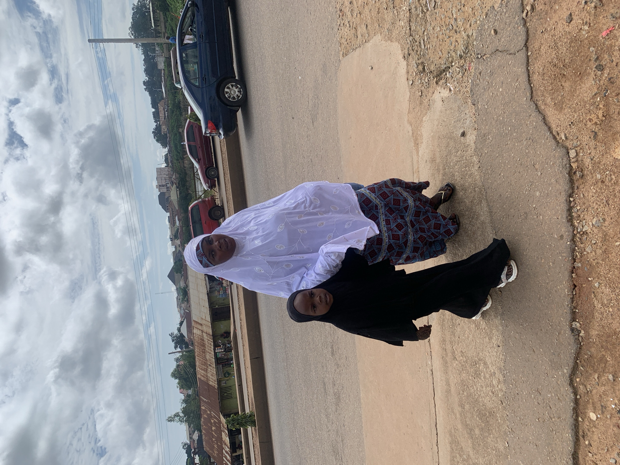 A woman in a white hijab and a child in a black hijab are walking on a dusty street in Bukuru, Nigeria. The woman is wearing a patterned skirt and white sandals, and the child is wearing black sandals. They are walking on a concrete sidewalk next to a road with cars driving by. In the background, there are buildings with corrugated metal roofs and trees. The sky is cloudy.