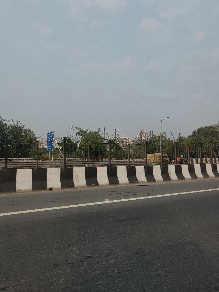 This image presents an outdoor scene captured from an elevated perspective, likely from a moving vehicle, on an overcast or hazy day in Ghaziabad, India. The foreground is dominated by a multi-lane asphalt road, featuring a continuous white lane marker. To the left of the road is a concrete barrier, distinctly painted with alternating vertical black and white stripes.
Beyond this barrier, an ornate black metal fence, adorned with decorative scrollwork along its top, extends horizontally across the frame. Behind the fence, a dense line of green trees and shrubs partially obscures a lower-level area, possibly another road or service lane. Through the foliage and fence on the left side, a blue rectangular road sign with white Hindi text is visible, although largely unreadable. It appears to be a directional sign, possibly indicating "Ghaziabad" and a distance such as "2km" or "20km."
Towards the right, a street light pole with a modern light fixture rises above the fence line. Directly beneath it, on the lower level, a yellow auto-rickshaw is clearly visible, either parked or stopped. Around the auto-rickshaw, several indistinct figures, likely people, and possibly other small vehicles like motorcycles, are gathered. In the distant background, through the hazy atmosphere and gaps in the trees, multiple residential or commercial buildings can be seen, characteristic of an urban or suburban environment. The sky is a uniform, pale blue-gray, indicating an absence of direct sunlight. No specific dynamic activity or event is taking place; the scene depicts a static moment along a typical Indian roadway.