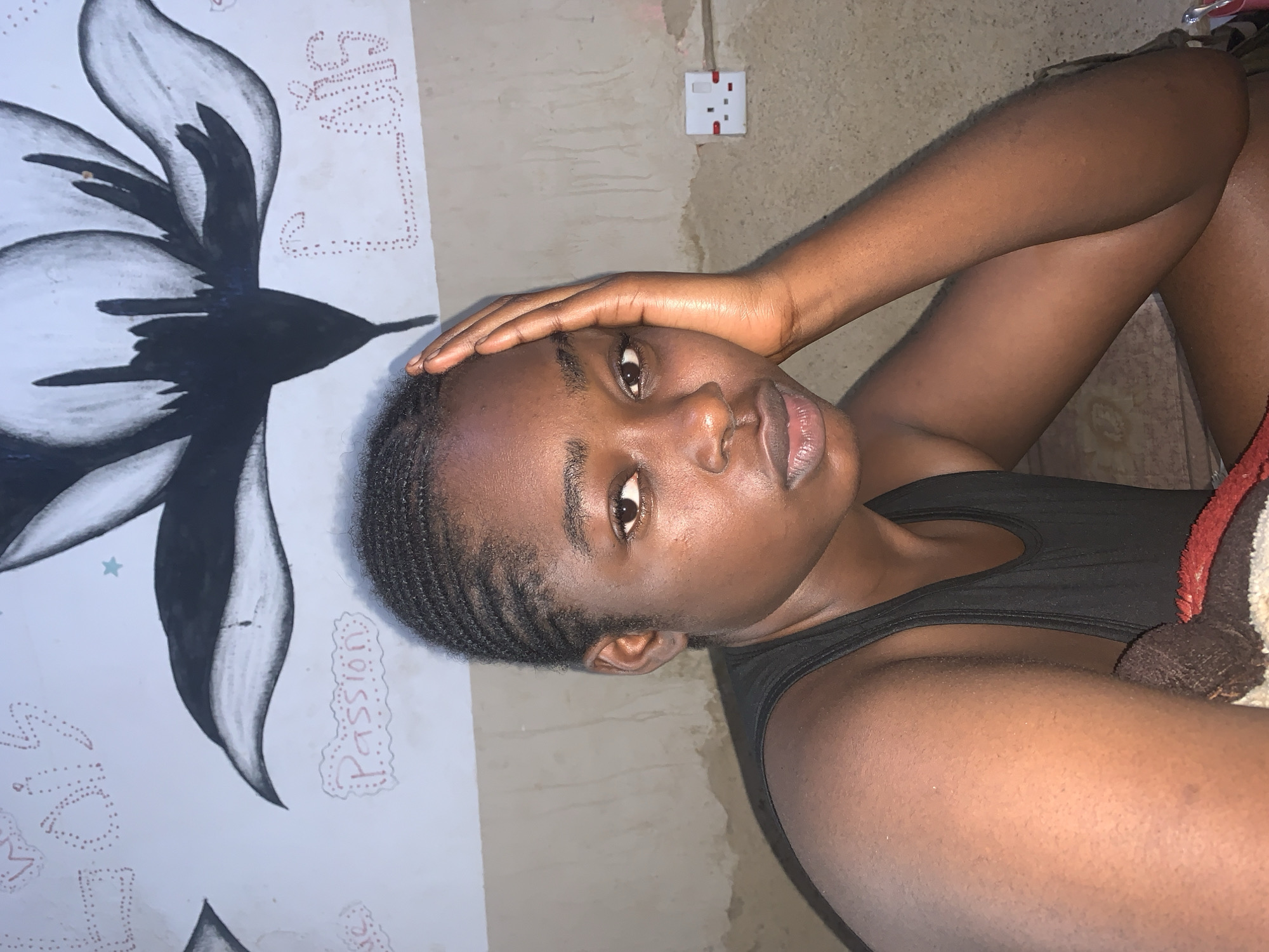 A medium shot captures a young Black woman indoors, seated with her right hand supporting her face. She is looking directly at the camera, her expression neutral. Her hair is styled in thin braids. She wears a black tank top. The background features a wall with a large black and white floral mural. To the right, a white electrical outlet with red accents is visible on the wall. The lighting is dim, and the overall impression is informal. The location is identified as Rafin Par, Nigeria.