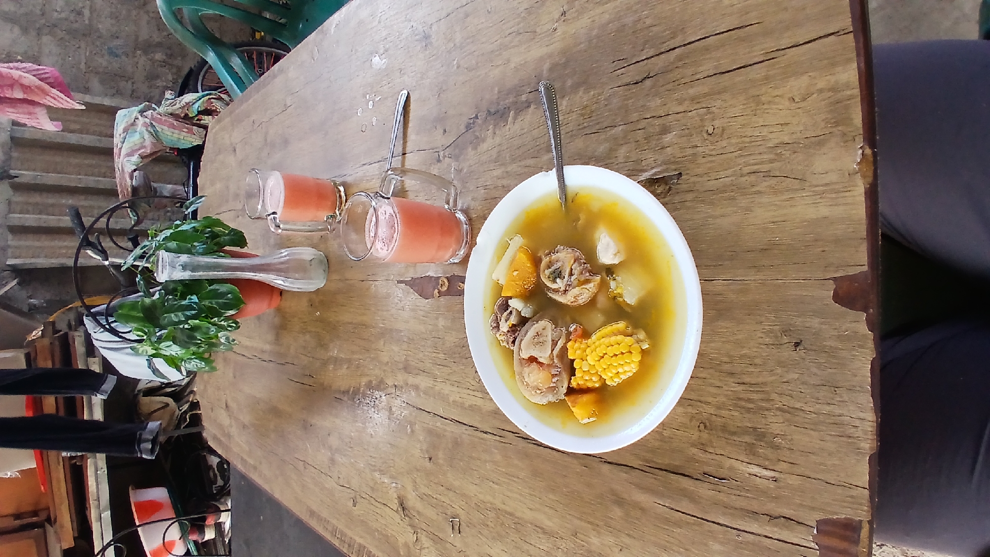On a weathered wooden table, potentially outdoors given the background elements, a white bowl contains a broth-based soup. The soup includes identifiable ingredients such as corn on the cob, chunks of squash, and bone-in meat. A spoon rests inside the bowl. Next to the bowl, two glass mugs filled with an opaque orange liquid, possibly juice or a smoothie, sit on the table. Further away, in a small planter with a clear glass insert, a leafy plant is visible. The overall scene suggests a meal is being served in what seems to be a casual setting. There is no strong evidence of a specific city, such as Altagracia, Venezuela.