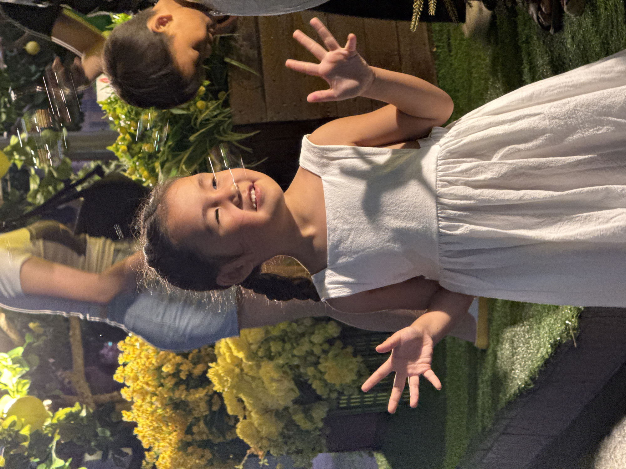 An outdoor photograph captures a joyful young Asian girl in what appears to be an evening or nighttime setting in Nha Trang Ward, Vietnam. The girl, with dark hair styled in a braid, is wearing a white sleeveless dress. She smiles broadly, looking upwards with her head tilted, and has both hands raised with fingers spread, as if interacting playfully with something in the air.
Numerous luminous, vertical streaks, resembling bubbles or water droplets, are suspended around her, catching the light and suggesting a moment of play or celebration. The background features a vibrant display of yellow flowers and lush green foliage, likely arranged in planters or garden beds. A section of artificial green grass is visible to the right. Blurred figures of other individuals, including another child, are partially visible in the background, hinting at a larger gathering or event.
The artificial lighting illuminates the scene, enhancing the festive atmosphere. The girl's expression radiates happiness and excitement. There is no visible text in the image. The overall scene conveys a cheerful outdoor event, possibly a garden party or festival.