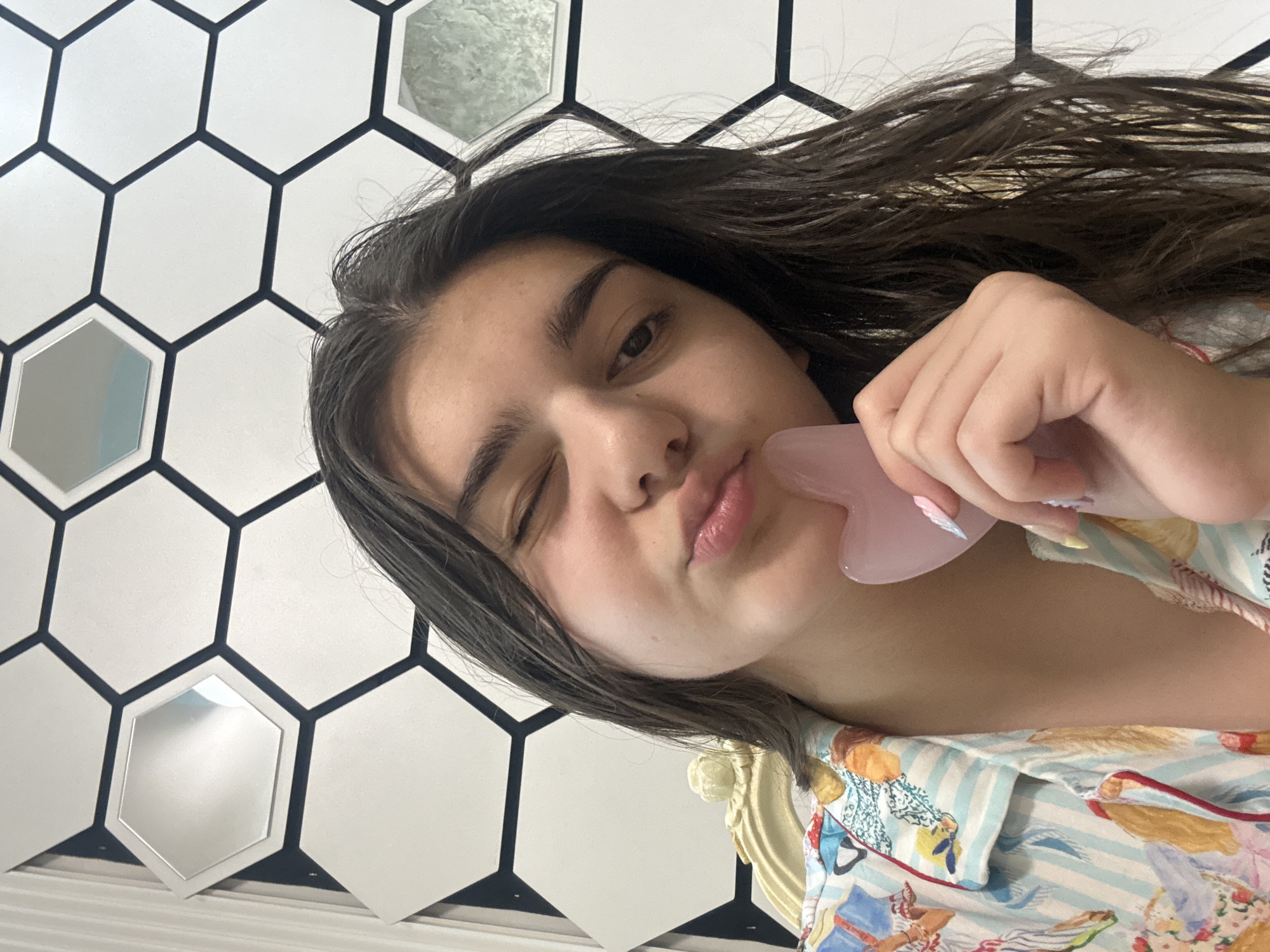 A young woman is holding a pink, heart-shaped object against her face. She is winking one eye and pursing her lips. She is wearing a printed, collared pajama top. Behind her is a wall with hexagonal tiles, some of which are mirrored. The location is Unknown, Unknown.