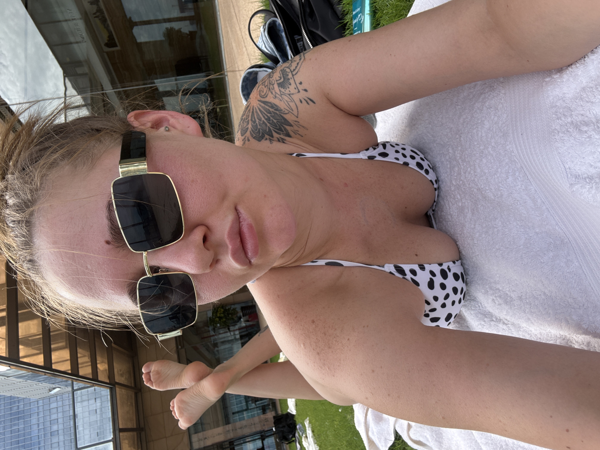 A woman is lying on a towel on grass, facing the camera. She is wearing a black and white polka dot bikini top and large, square, gold-rimmed sunglasses. Her hair is pulled back. Behind her, a building with reflective windows and a modern architectural design can be seen, along with a section of a glass structure with orange supports. The scene appears to be in an outdoor urban setting, possibly a hotel courtyard or public area in Yiwu, China, given the architectural style. There is a black bag and some other items on the grass to her right. Her feet are visible in the upper left of the frame, positioned at an angle. She has a tattoo on her left shoulder. The lighting suggests it is daytime.