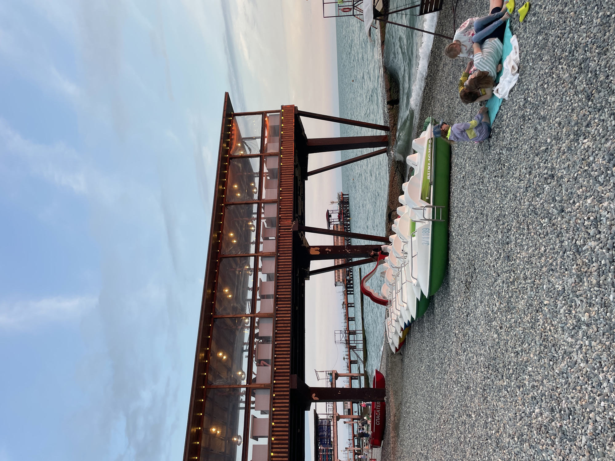 The image depicts a rocky beach in Sochi, Russia, with a pier and an elevated restaurant structure in the background. In the foreground, a line of paddle boats is parked on the beach. Several people are seated on a blanket near the paddle boats, including two children and an adult. The sky is partly cloudy, and the sea is calm with gentle waves. The lighting suggests it is either early morning or late evening. The restaurant has a covered outdoor seating area with lights illuminating the tables. There is also a red boat labeled "СПАСАТЕЛЬ" (Lifeguard) on the beach.