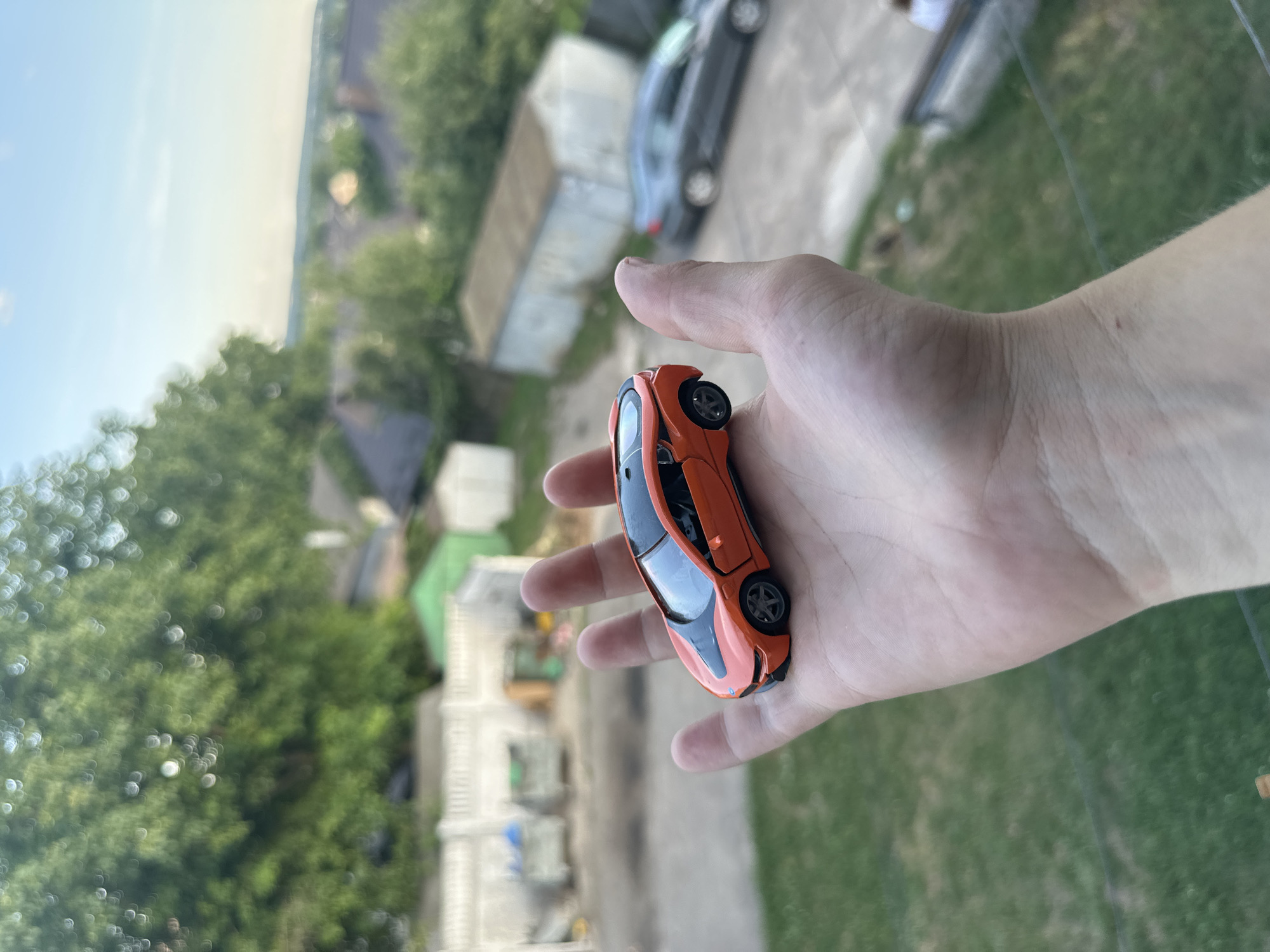 A first-person perspective shot shows a hand holding an orange toy car with a black roof and hood, placed on the palm. The background, slightly out of focus, reveals a suburban or rural setting. A white fence runs horizontally across the middle ground, with dumpsters visible behind it. Further in the background, several houses and garages are scattered among trees and green grass under a pale sky. The scene appears to be in a residential area with a slightly overgrown feel.