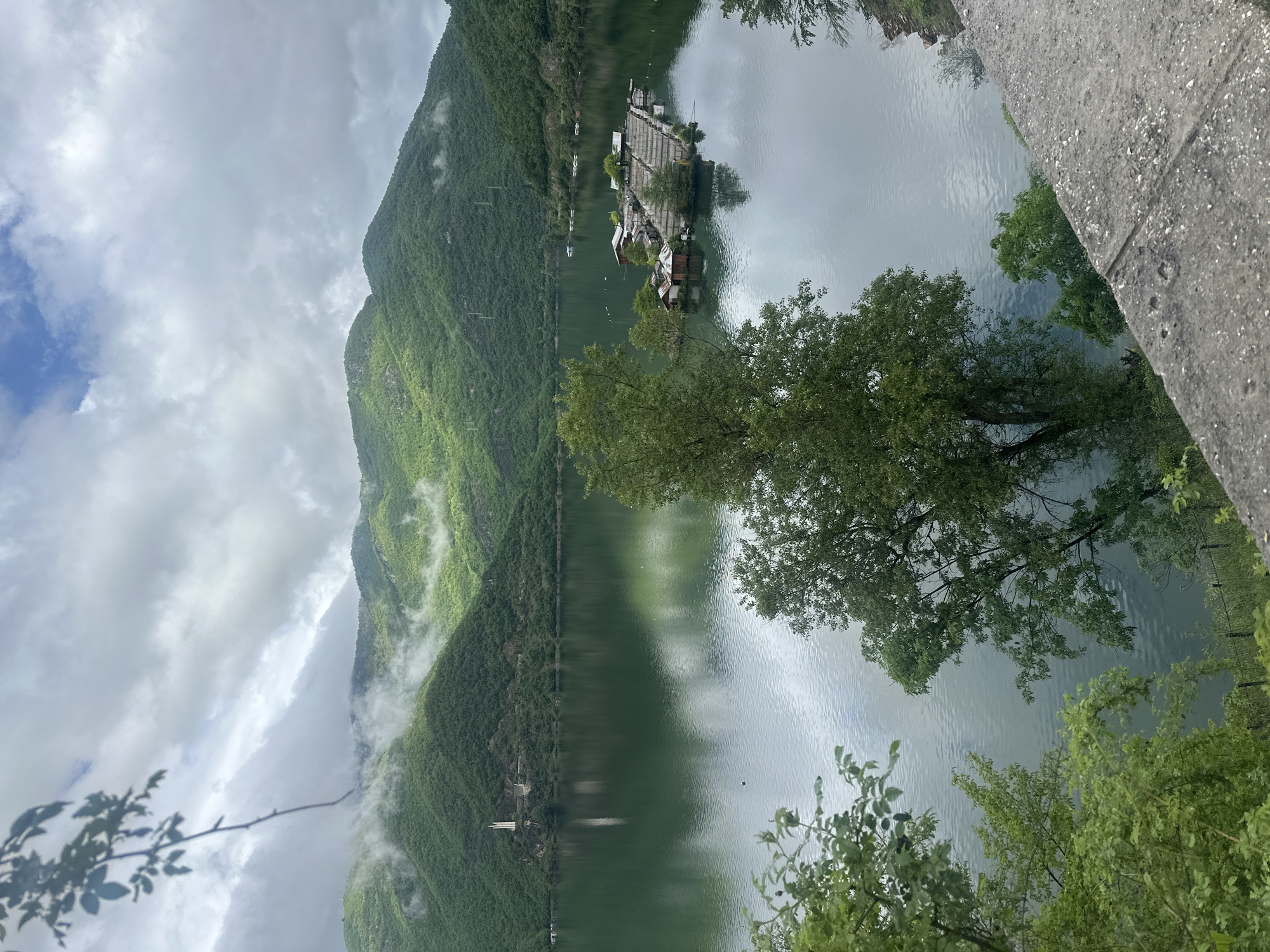 The image depicts a mountainous landscape with a body of water, possibly a lake or reservoir, in the foreground. Lush green vegetation covers the mountainside, with some areas obscured by low-lying clouds or mist. In the water, there is a cluster of structures, possibly floating platforms or houseboats. Trees and vegetation frame the view from the foreground, adding depth to the composition. The photo seems to be taken from an elevated position, such as a bridge or hillside. Based on the prompt's information, the location is Mihalkovo, Bulgaria.