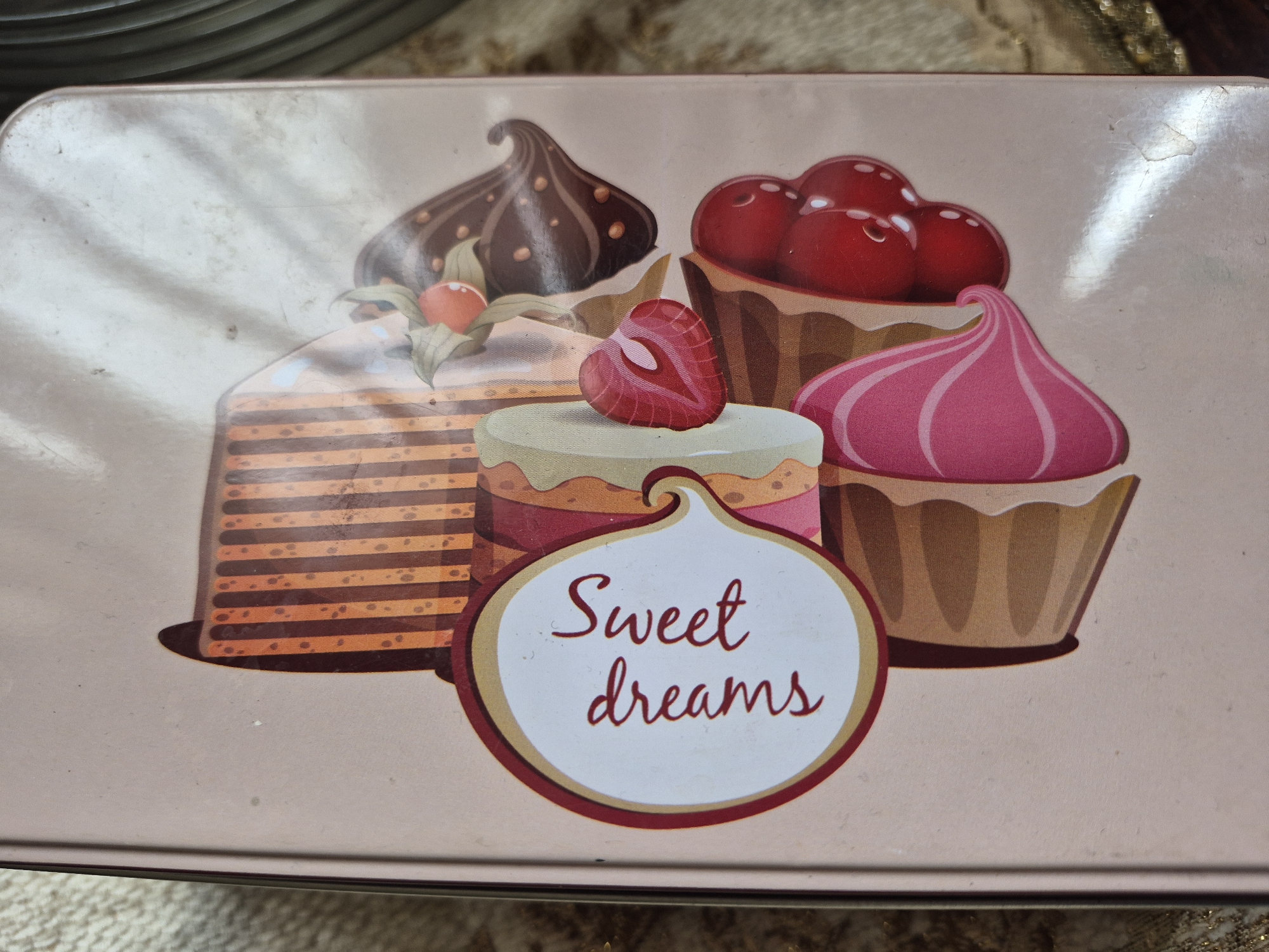 The image shows a close-up view of a light pink tin with a decorative illustration of desserts. The illustration features stylized cakes and pastries, including what appears to be a strawberry, cherries, and whipped cream. A speech bubble-like element contains the words "Sweet dreams" written in a cursive, reddish-brown font. The tin has a slightly worn appearance, with some smudges and scuffs visible on its surface. The background of the image is a textured, beige material. The image does not provide any information about the city of Vanadzor, Armenia, or any people or actions taking place.