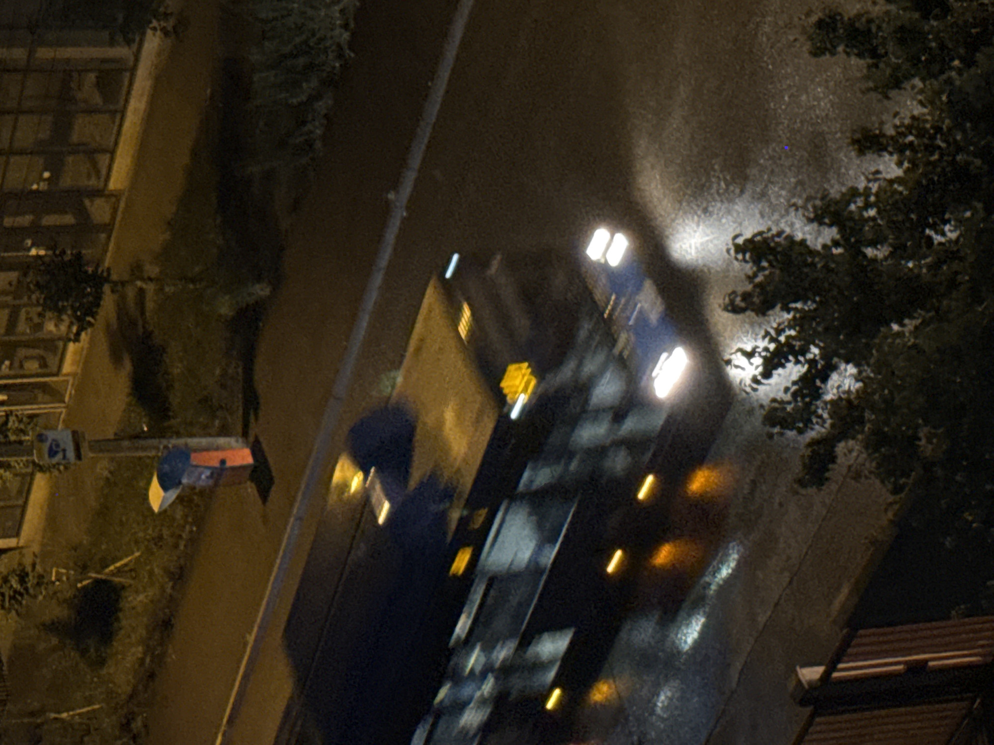 An out-of-focus, nighttime exterior shot depicts a bus moving on a wet street. The bus is dark with yellow and white lights visible. The street reflects the lights of the bus and other unseen sources. In the background, a building with large windows and a street lamp are visible. To the left, a bus stop shelter with a red and yellow sign is partially obscured by the bus. Trees and other foliage are present in the foreground and background, suggesting an urban environment. The image does not provide enough detail to determine the specific location in Tbilisi, Georgia.