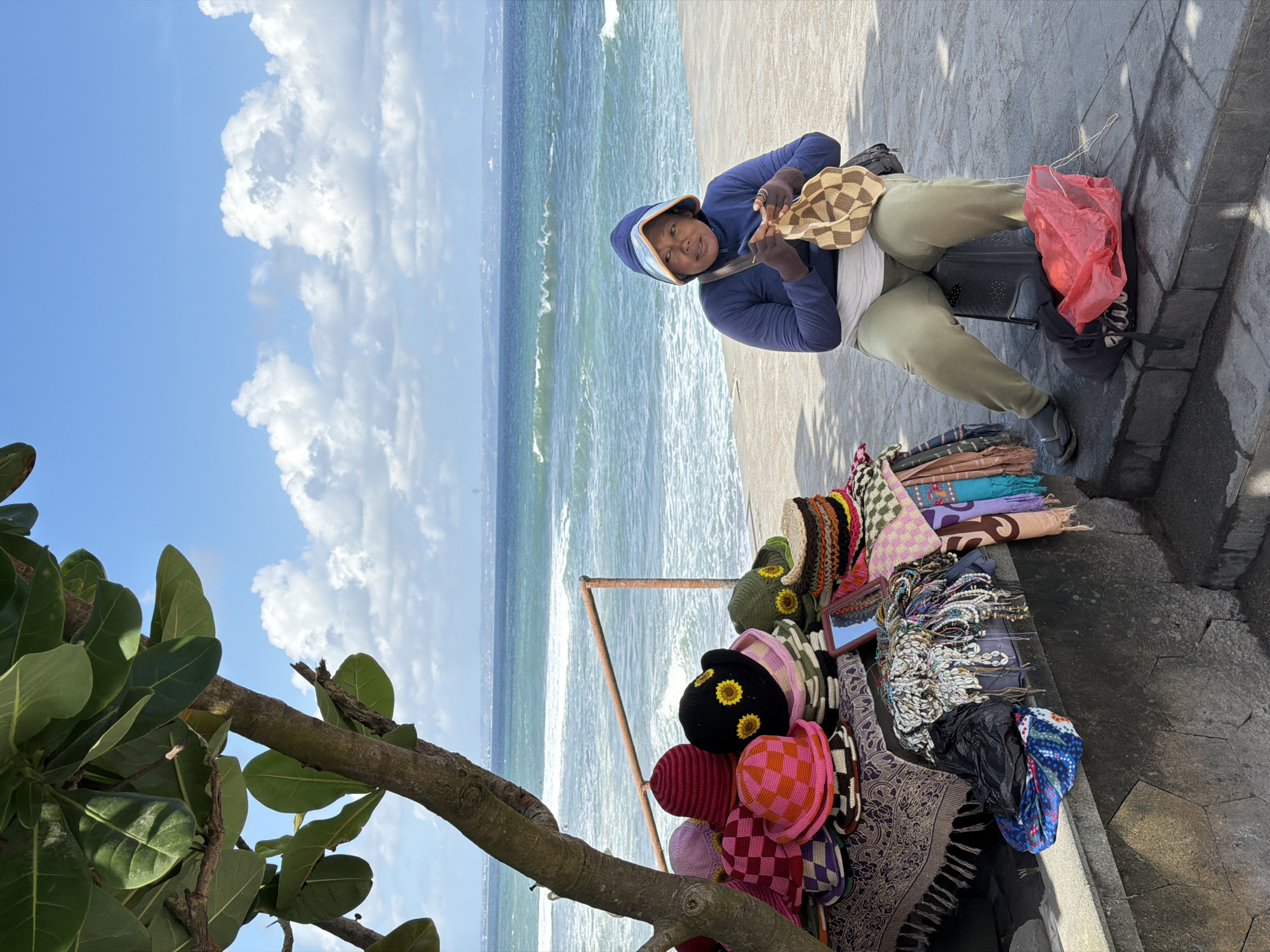 A woman sits on a stool in front of a table of hats and scarves, knitting. The scene is outdoors, with the ocean and a blue sky with clouds in the background. The woman is wearing a blue hooded jacket and light green pants. She is holding a checkered bag and knitting. The table is covered with a patterned cloth and is laden with various knitted hats, scarves, and beaded necklaces. A large tree with green leaves is to the left of the table. The ground is a concrete walkway. The location appears to be a beachside vendor stall. The image is likely taken in Mengwi, Indonesia, given the context of beachside vendors and the style of goods.