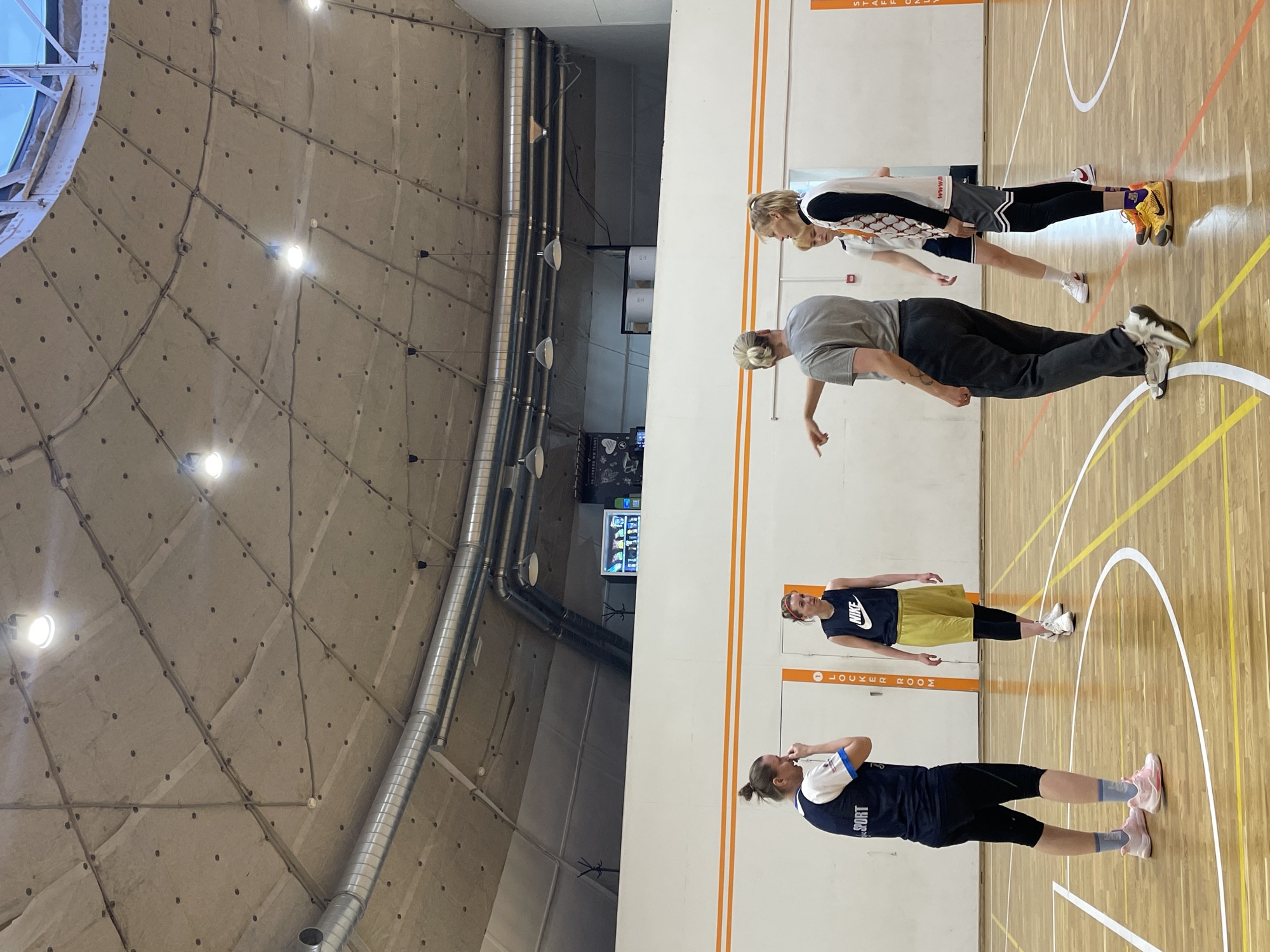 The image depicts an indoor basketball court in what appears to be Saint Petersburg, Russia, based on architectural and linguistic cues. Four women are present. One wears a dark blue shirt with the word "SPORT" on it and black tights, and pink running shoes. Another woman wears a black Nike tank top and light-colored shorts with white sneakers. A third person is wearing a grey t-shirt and dark pants and white sneakers, pointing. The fourth woman is wearing a basketball jersey with shorts and yellow basketball shoes. The court has standard basketball court markings in white, yellow, and orange. In the background, there is a sign labeled "LOCKER ROOM." The building has a distinctive dome-shaped ceiling. Exposed pipes run along the upper left. A vending machine is visible in the background.