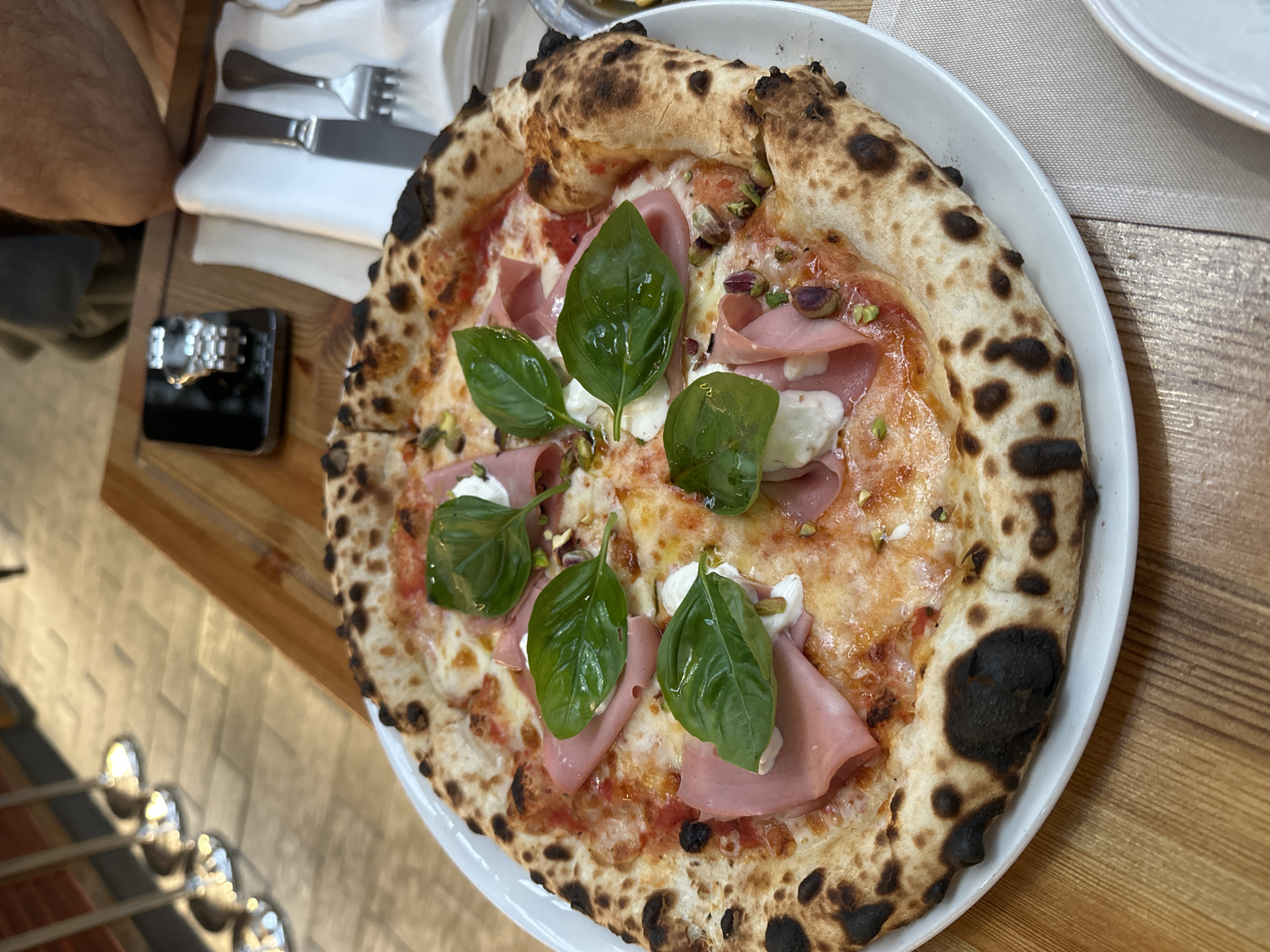 The image depicts a pizza on a white plate, placed on a wooden table. The pizza is topped with ham, cheese, basil leaves, and appears to have pistachios scattered on top. The crust is charred in places. To the left of the plate are silverware and what appears to be a watch or phone on a wooden surface. In the background, additional silverware is visible. The scene is likely indoors, possibly in a restaurant setting in Yerevan, Armenia. There is a person partially visible at the top left corner of the frame.