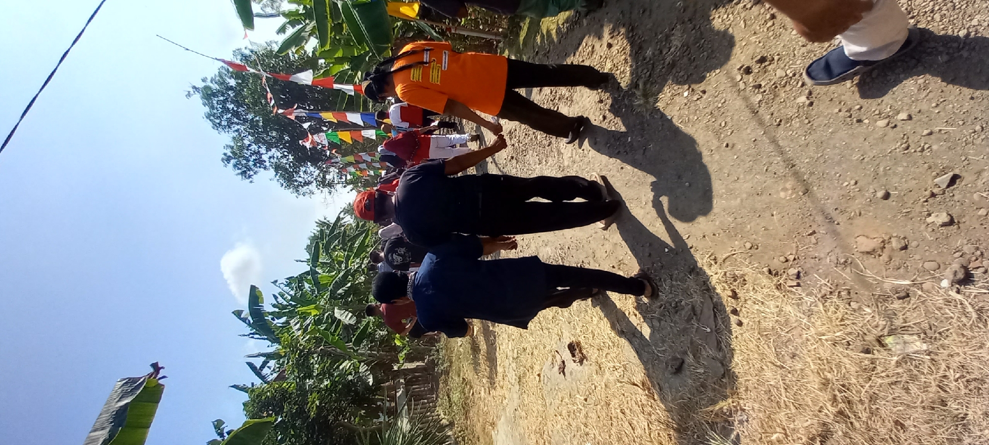 In Sidamulya, Indonesia, on a dirt road bordered by banana trees, a group of people walk away from the camera. Three individuals are in the foreground, with two walking hand-in-hand. The person on the left wears a dark blue jacket and dark pants, while the person next to them wears a black t-shirt and black pants. The person on the far right is wearing an orange t-shirt with writing on the back and dark pants. They are walking on a dusty, gravel path with dry grass along the side. In the background, other people are visible, as well as a line of colorful flags strung between trees. The sky is clear and blue with a few small clouds, suggesting it is daytime and sunny.