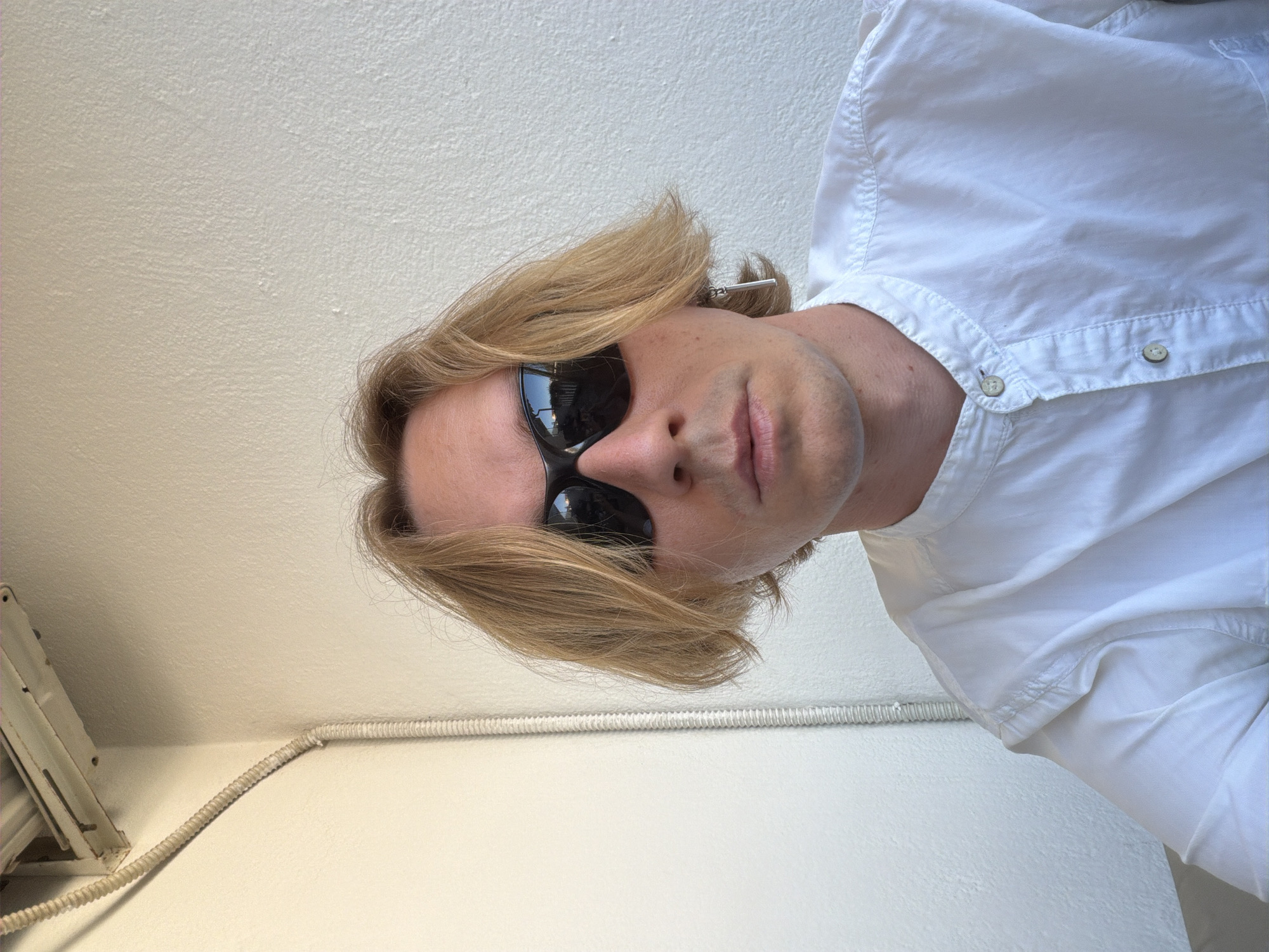 The image shows a low-angle, close-up shot of a person wearing a white button-up shirt and black sunglasses. The person has blonde hair, which is styled to one side. The background is a textured, light-colored wall, possibly a ceiling, with some electrical conduit visible along the bottom. The lighting suggests it might be daytime, possibly indoors or in a shaded outdoor area, as there are no strong shadows. The overall mood is casual and perhaps a bit relaxed. No text is visible in the image. The provided location context of Budva, Montenegro, doesn't offer specific visual cues within the image itself, but it sets a general geographical context.