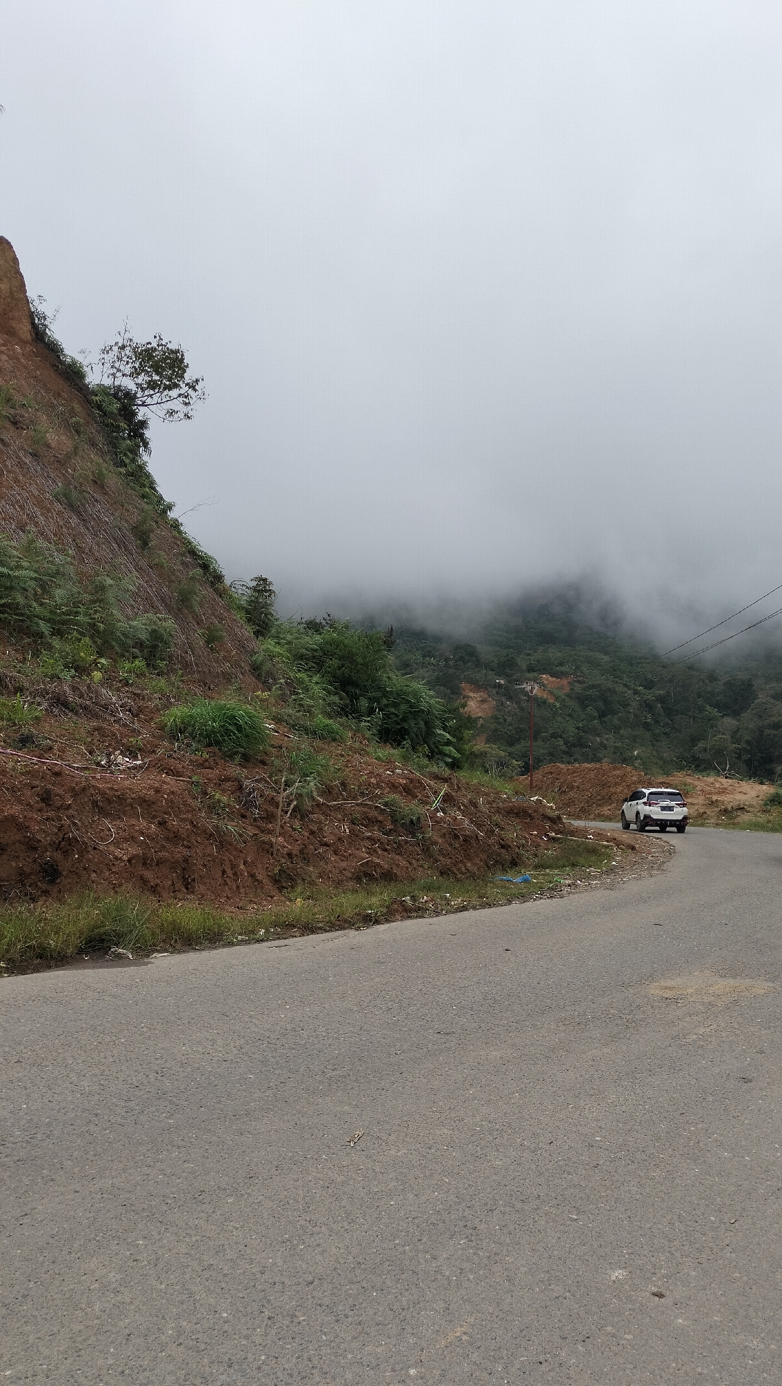 The image depicts a scenic view of a winding road passing through a mountainous, forested area with a light mist or fog. On the left, a steep, eroded slope covered with brown soil and sparse vegetation rises above the road. The road itself is paved and appears to be in good condition. In the middle ground, a white SUV is parked on the side of the road. Beyond the road, a lush green forest stretches across the hills, partially obscured by a soft, grey mist that also blankets the distant mountain peaks. Utility poles with wires are visible on the right side of the image, suggesting proximity to some form of infrastructure. The overall atmosphere is serene and natural, with a sense of elevation and expansive views.