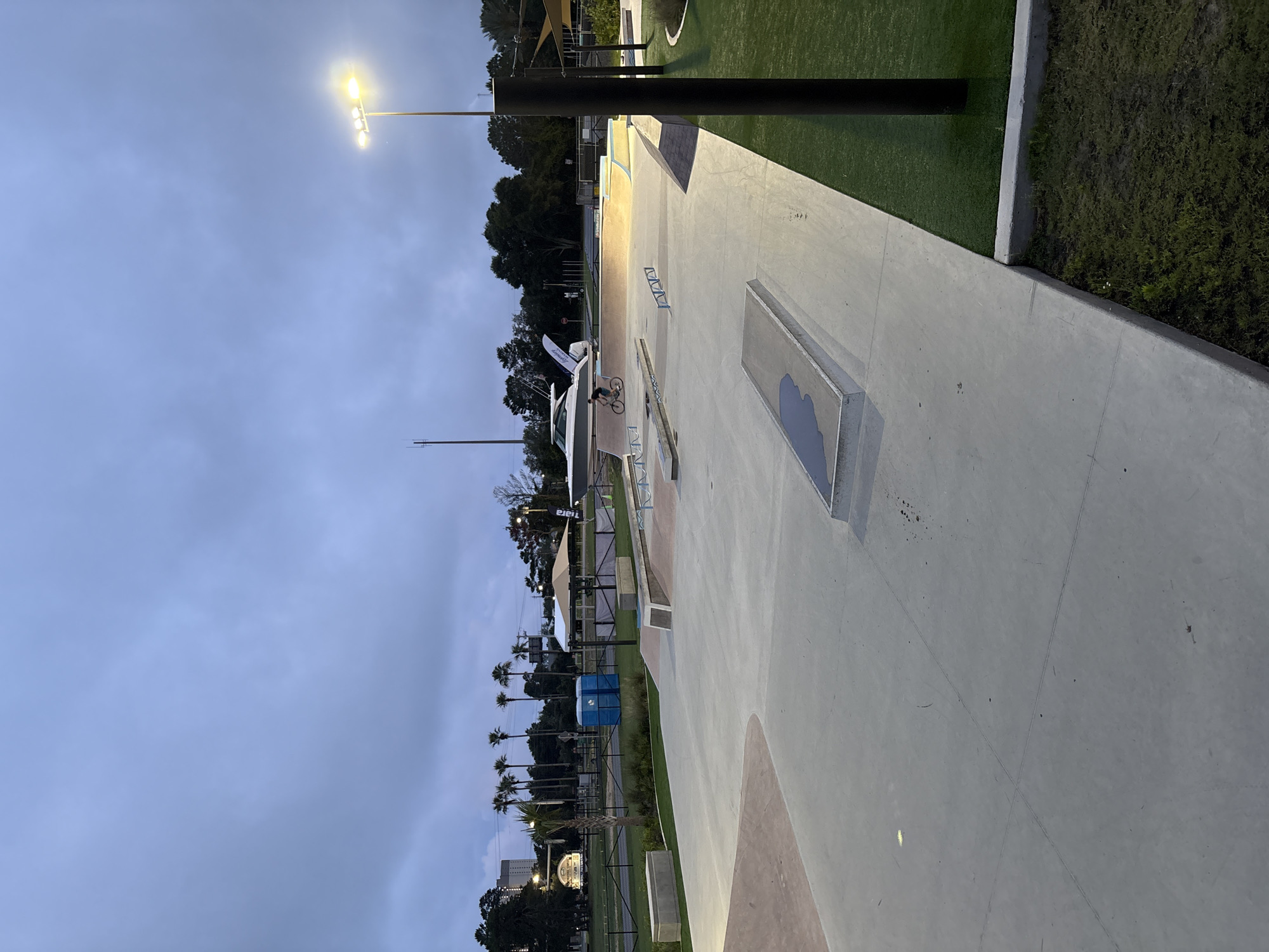 This image shows a skate park at night, likely in Panama City Beach, Florida, based on the vegetation and surroundings. The skate park is constructed of concrete with various ramps, rails, and bowls. An individual is on a bicycle in the middle of the skate park. Artificial turf and landscaping borders the concrete skating area. A tall pole with lights illuminates the park. The sky is overcast. Palm trees are visible in the background. Blue portable toilets and tents are situated in the distance beyond the skate park features.