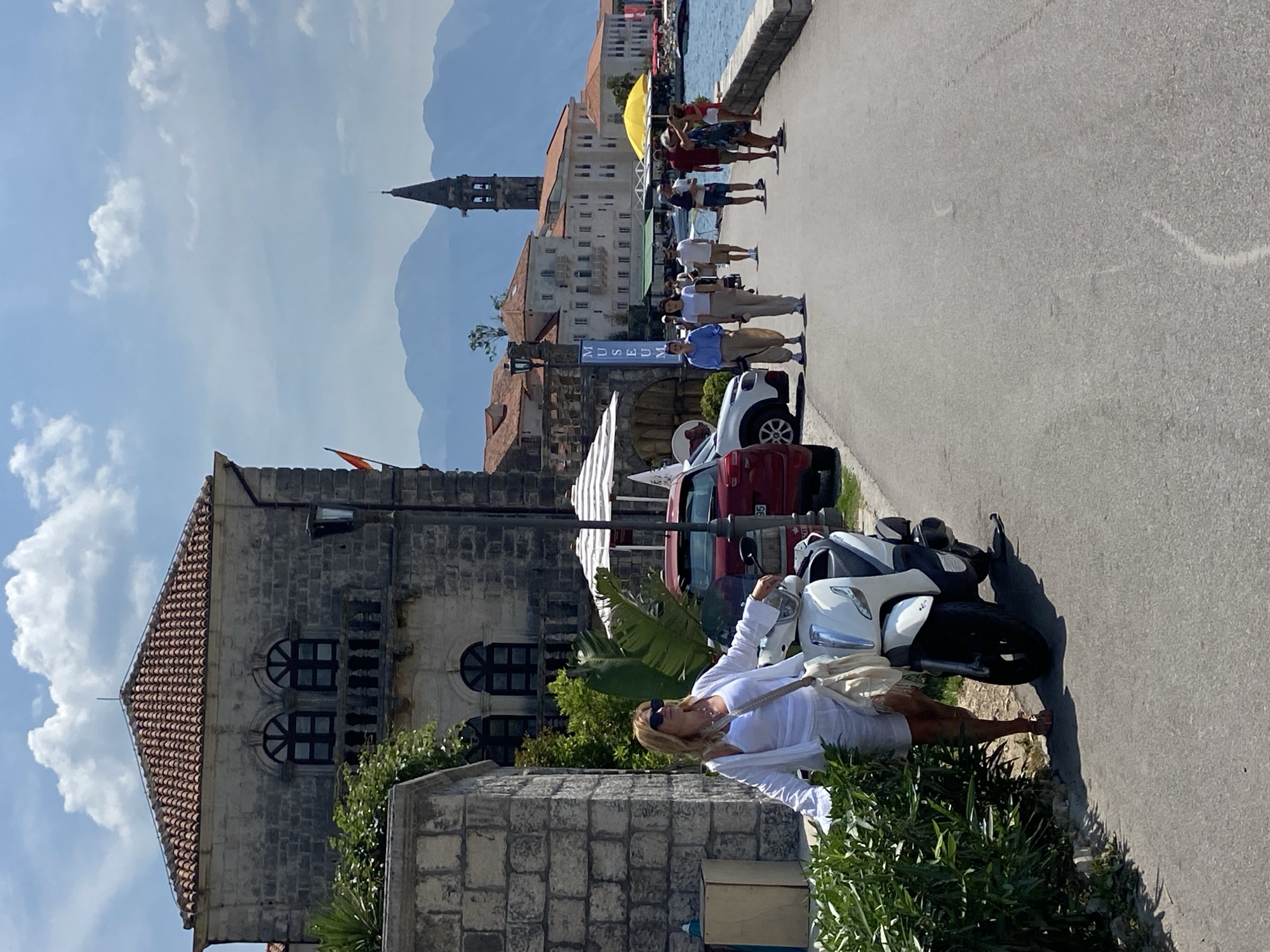 In Perast, Montenegro, a woman stands next to a white scooter on a sun-drenched promenade. She is wearing white linen clothing and a straw hat, and she is looking to her right. Behind her, a stone building with arched windows and a red-tiled roof is visible. In the background, several people walk along the promenade, and beyond them, the blue waters of the bay are dotted with boats. Mountains form a majestic backdrop to the scene under a partly cloudy sky. The architecture suggests a historic European coastal town.