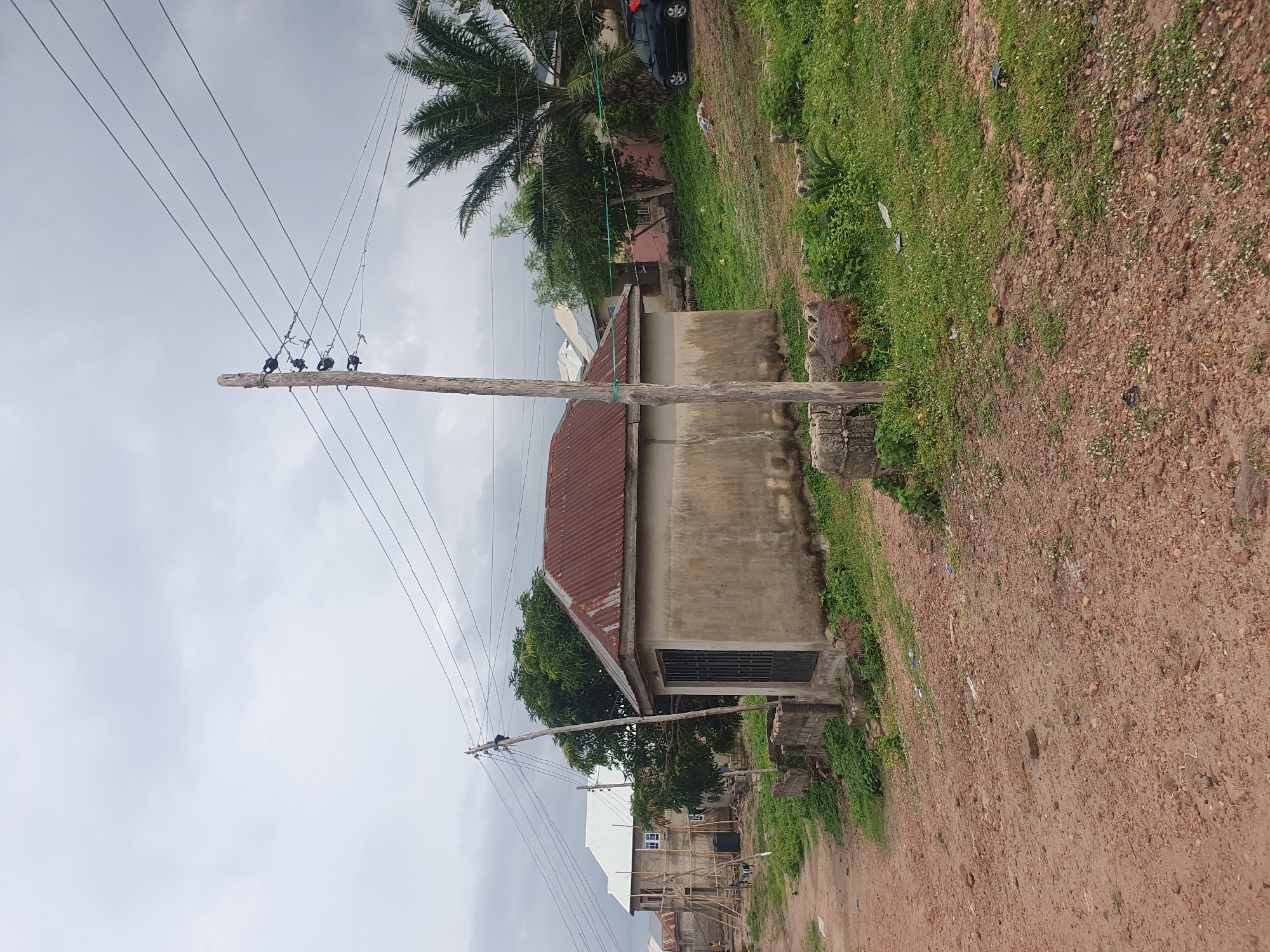 The image depicts a somewhat overcast day in Lokoja, Nigeria, with a focus on a small, concrete building with a corrugated iron roof. The building has a metal gate on its front. A wooden utility pole stands to the right of the building, supporting multiple overhead electrical wires that stretch across the frame. In the background, to the left, a multi-story building with a white roof is under construction, supported by scaffolding. Several trees are visible, including a palm tree on the right side of the image. A dark-colored car is parked on a grassy area behind the concrete building. The ground in the foreground is a dirt road with scattered stones and small patches of grass and weeds.