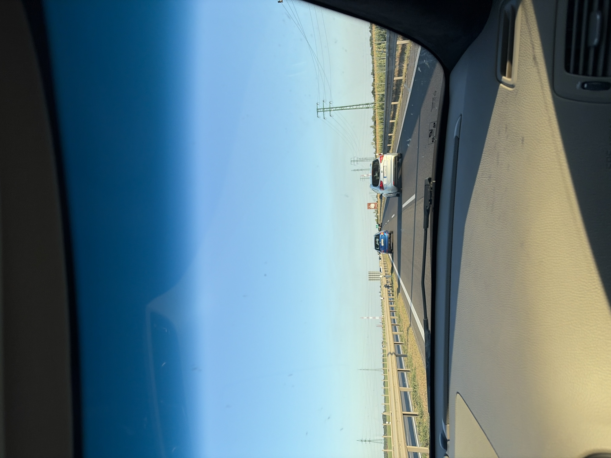 The image is taken from the interior of a vehicle, looking forward through the windshield. The sky is a clear, vibrant blue, transitioning to a deeper blue near the top edge of the frame, suggesting a sunny day. The dashboard of the car, a textured beige material, is visible in the foreground, casting a shadow. Outside the car, a highway stretches into the distance. Several cars are on the highway, including a blue car in the left lane and a silver car in the right lane, both moving away from the camera. A metal guardrail runs along the side of the highway. Beyond the guardrail, there are fields of crops, likely corn, and utility poles with power lines are visible against the horizon. The location is the highway near Križovany nad Dudváhom, Slovakia.