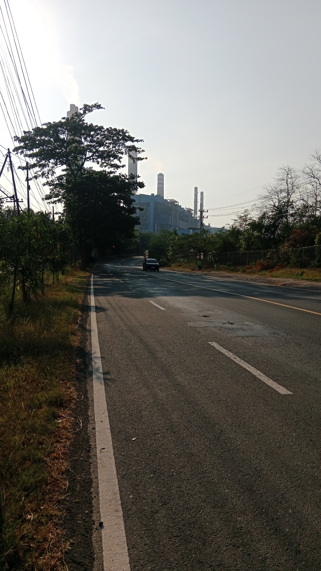 The image depicts a road leading towards a large industrial complex with multiple smokestacks, one of which is emitting a plume of white smoke. The complex appears to be a power plant. The road is asphalt, with white and yellow lane markings. On the left side of the road, there is a grassy verge with some trees and vegetation. Overhead power lines are visible against the sky, which is a pale, hazy blue. A dark-colored car is driving on the road in the distance. The environment suggests an industrial area on the outskirts of a city. The location is identified as Bhinar, Indonesia.