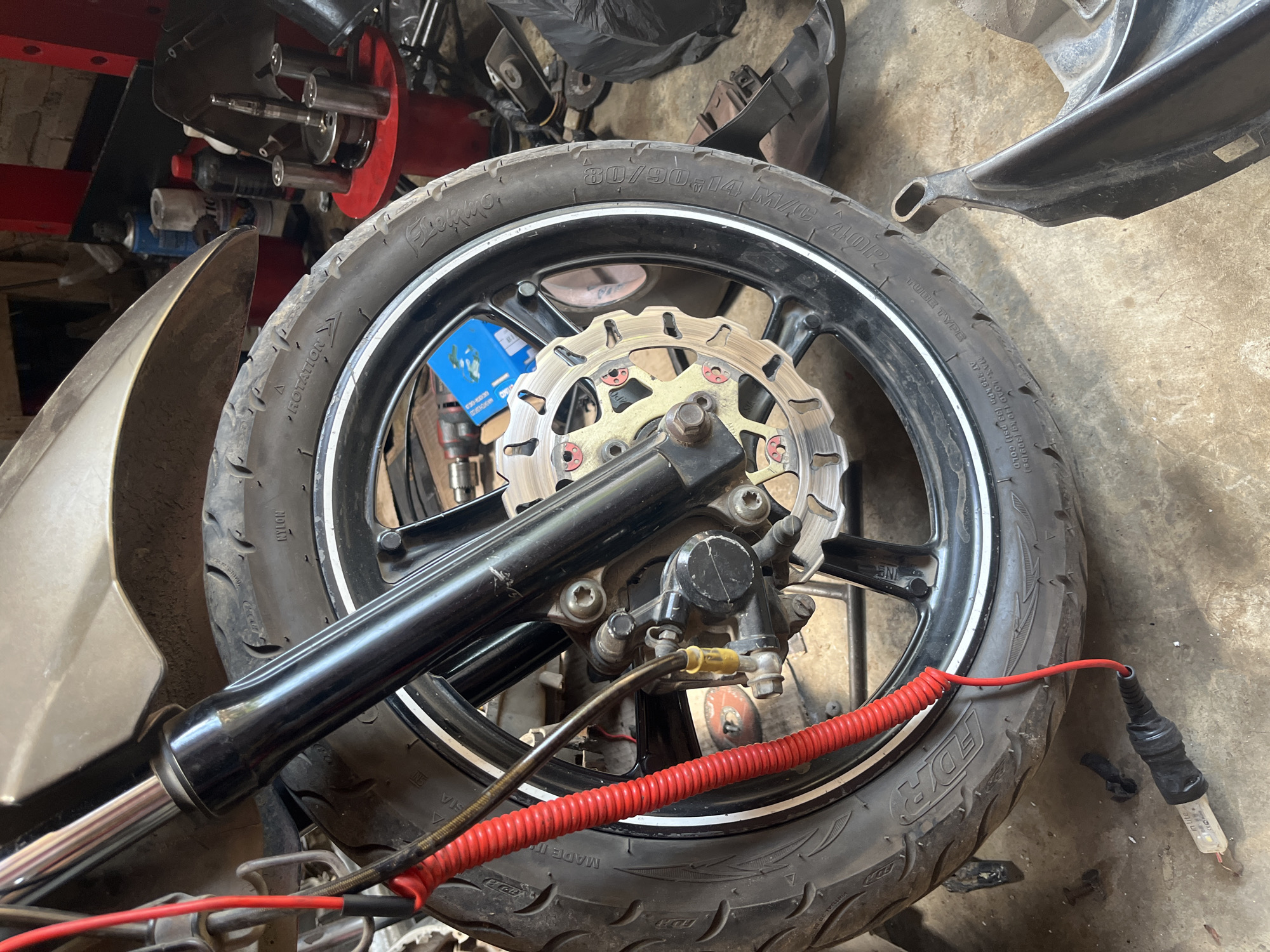 A close-up, ground-level shot captures the front wheel, suspension, and brake assembly of a motorcycle. The motorcycle's front tire, branded "FDR" and with tire size "80/90-14", is prominent. A shiny silver brake disc with gold-colored bolts is visible behind the tire, attached to the wheel hub. The black motorcycle fork is positioned above the wheel, with a bright red coiled wire running down alongside it. A black electrical cable with a clear glass bulb at the end is resting on the concrete floor in the foreground, connected to the red wire. The background is cluttered with various tools, spray cans, and motorcycle parts, suggesting a workshop or repair area in Kebumen, Indonesia. The lighting is dim and uneven.