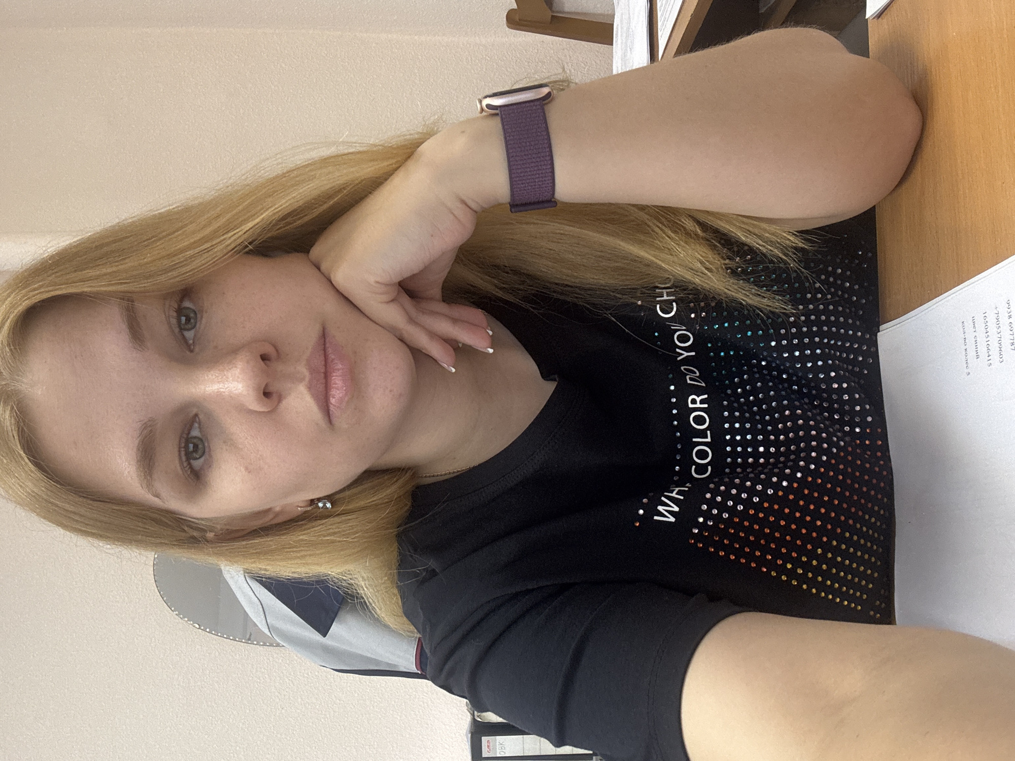 A woman with blonde hair rests her head on her hand, which is adorned with a purple smart watch. She is wearing a black t-shirt with the text "Wh. COLOR Do YOU CH" and a gradient of colored dots. She is sitting at a desk, which contains papers. The location is Rusanovo, Russia.