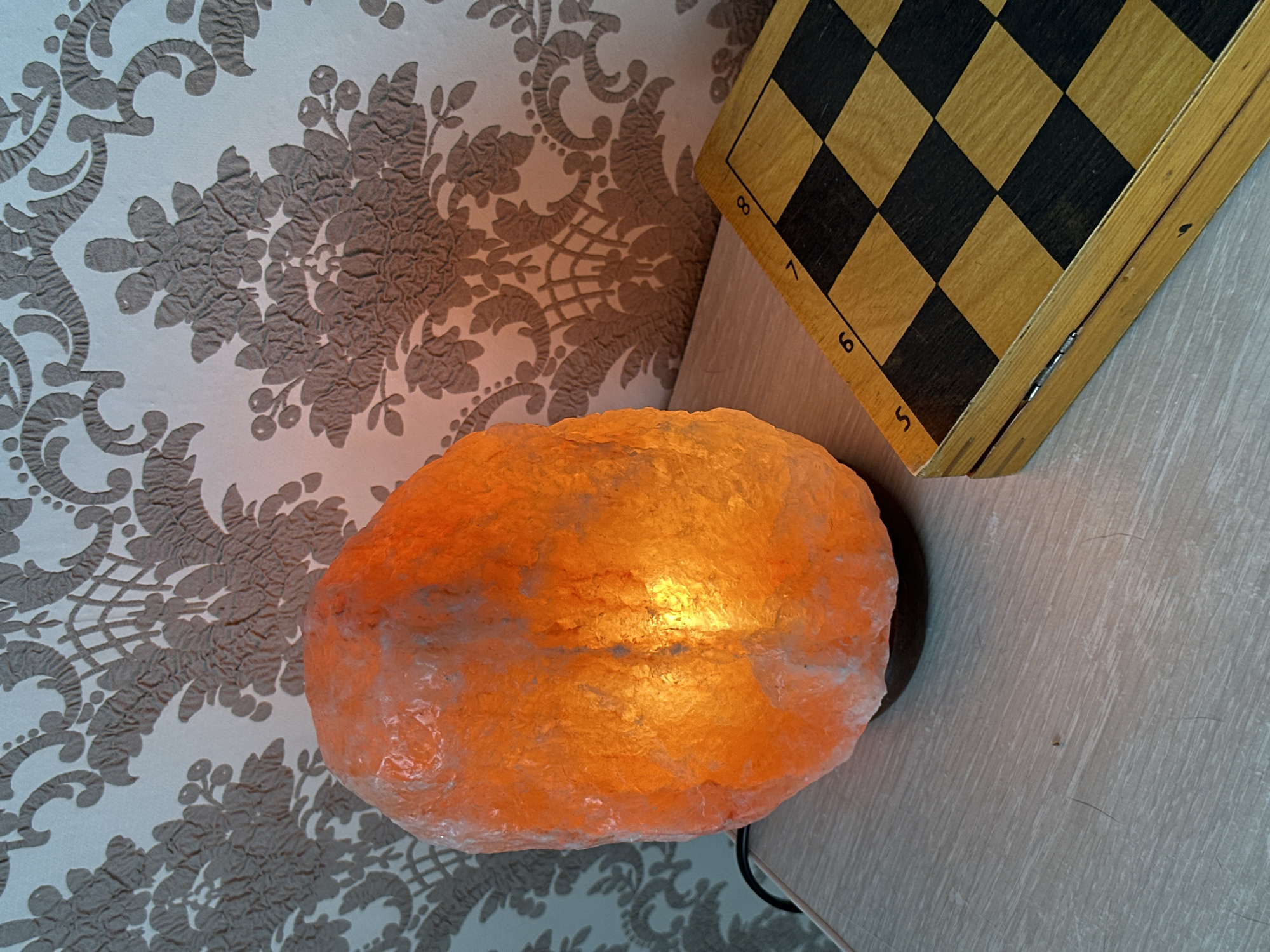 A Himalayan salt lamp, glowing with an orange light, sits on a light-colored wooden table next to a closed wooden chessboard. The lamp is irregular in shape and appears to be resting on a dark base. The chessboard is made of light and dark wood squares and has numbers etched along one edge, ranging from 5 to 8. Behind the lamp and chessboard is a textured wallpaper with a damask pattern in shades of grey and white. The overall scene suggests an indoor setting, possibly a home or a lounge area. There is no indication of the city of Energetik, Russia, in the image.
