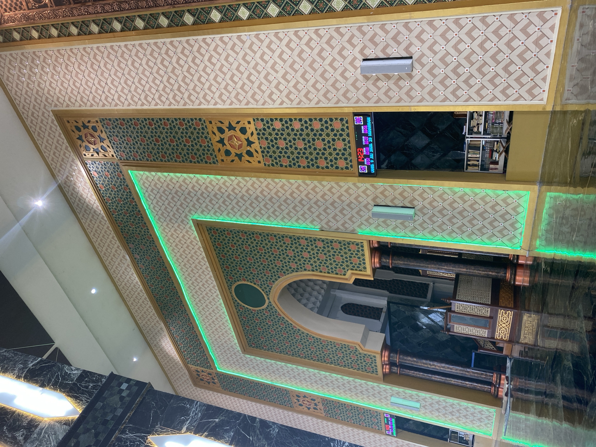 The image shows a section of the interior of a mosque, characterized by ornate Islamic geometric patterns and gold accents. The wall is decorated with a repeating pattern of beige diamond shapes with small red squares in their centers, framed by geometric borders in green and gold. A large, arched doorway is central to the image, adorned with intricate green and gold patterns. Green LED lights trace the edges of the doorway and the surrounding architectural features, creating a vibrant glow.
To the left of the doorway, a large marble-clad column with dark veining is visible, extending upwards towards the ceiling which features bright, rectangular light fixtures. To the right of the doorway, a digital display shows prayer times and dates. Below this display, a shelf unit filled with books is visible.
In the foreground, the highly polished floor reflects the architectural details and the green lighting. A wooden minbar, a raised pulpit, is positioned within the alcove of the doorway. Dark, decorative columns with copper-colored capitals support the upper structure. The overall impression is one of grandeur and meticulous craftsmanship. The location is identified as Aluekambuk, Indonesia.